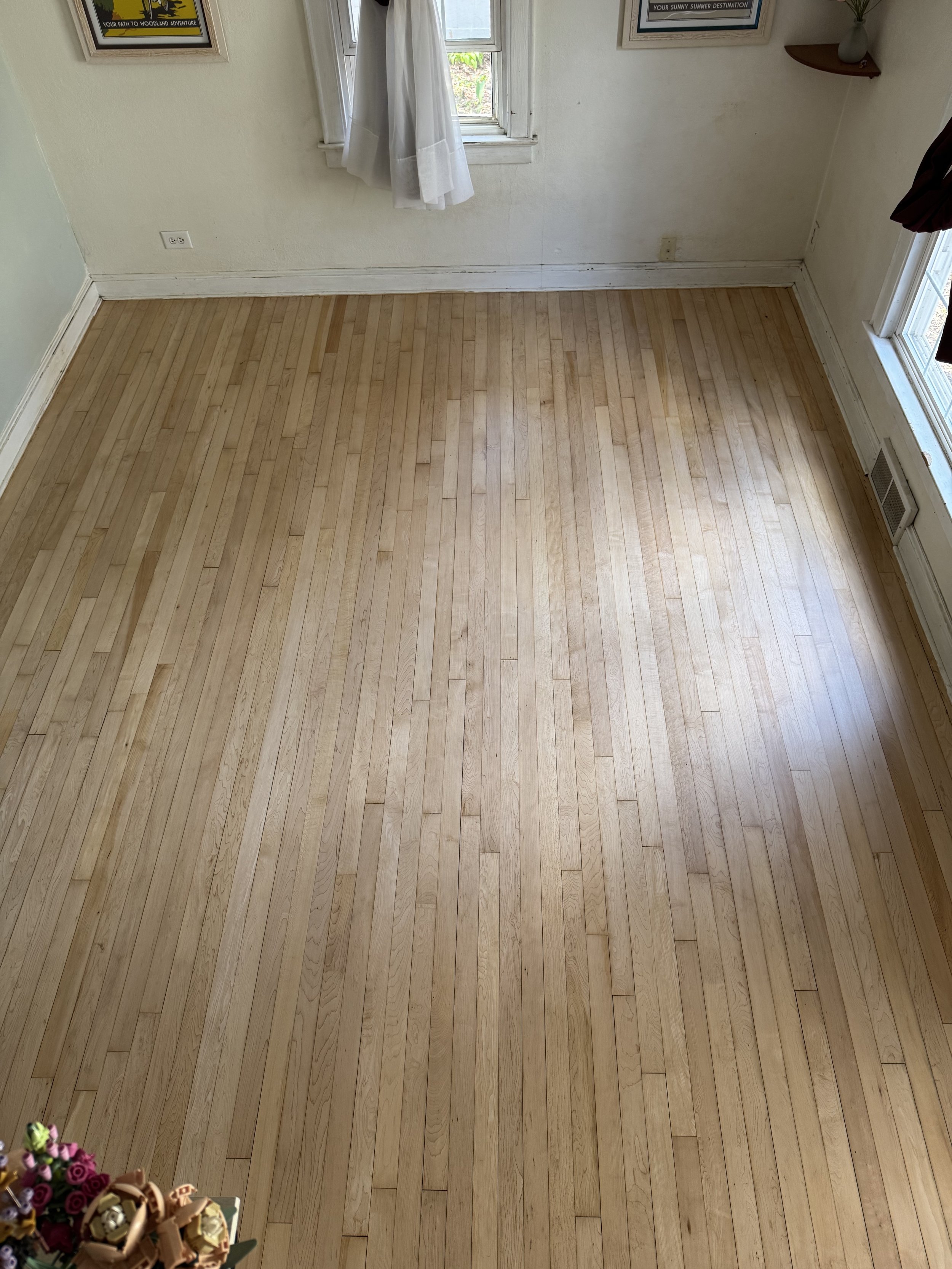 Empty room with light-colored wooden floor, white walls, a window with white curtains, and framed posters on the walls.