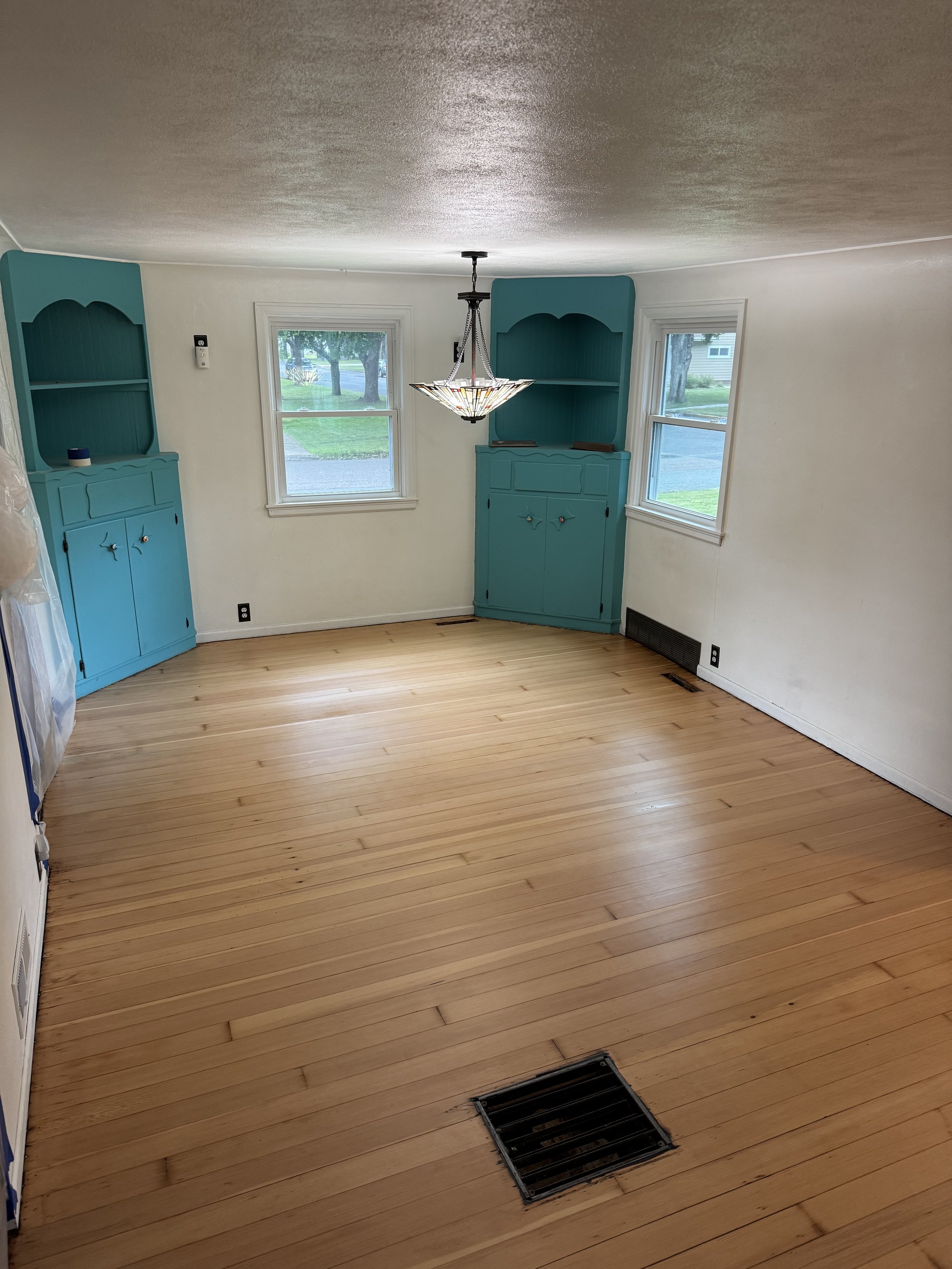 Empty living room with wooden floors, two windows, white walls, a chandelier, and two blue corner cabinets.