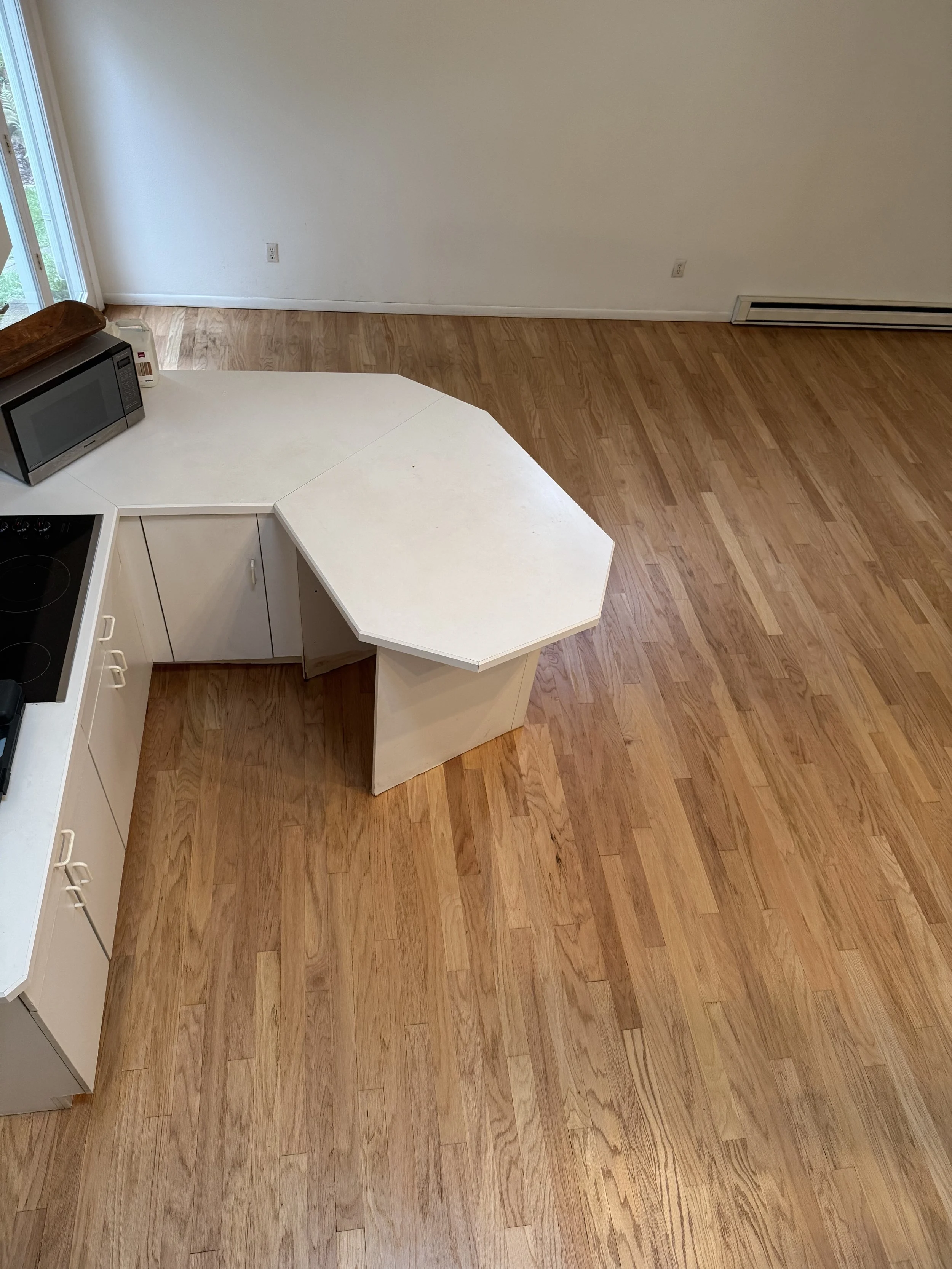 Empty kitchen with hardwood floors, white cabinetry, a white countertop with an octagonal extension, a microwave, and a stove.