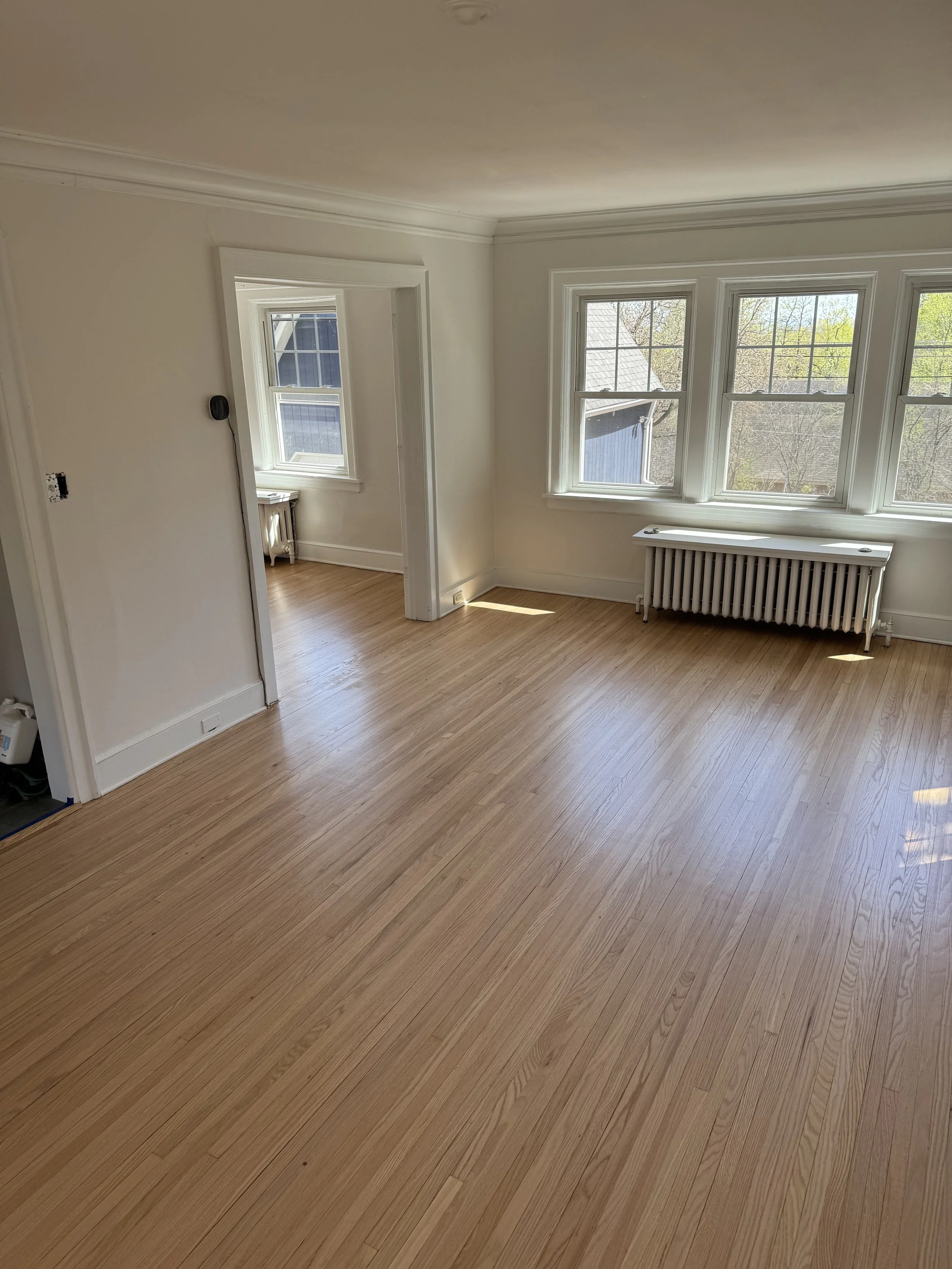 Empty room with light wood flooring, white walls, large windows, and a radiator underneath the windows.