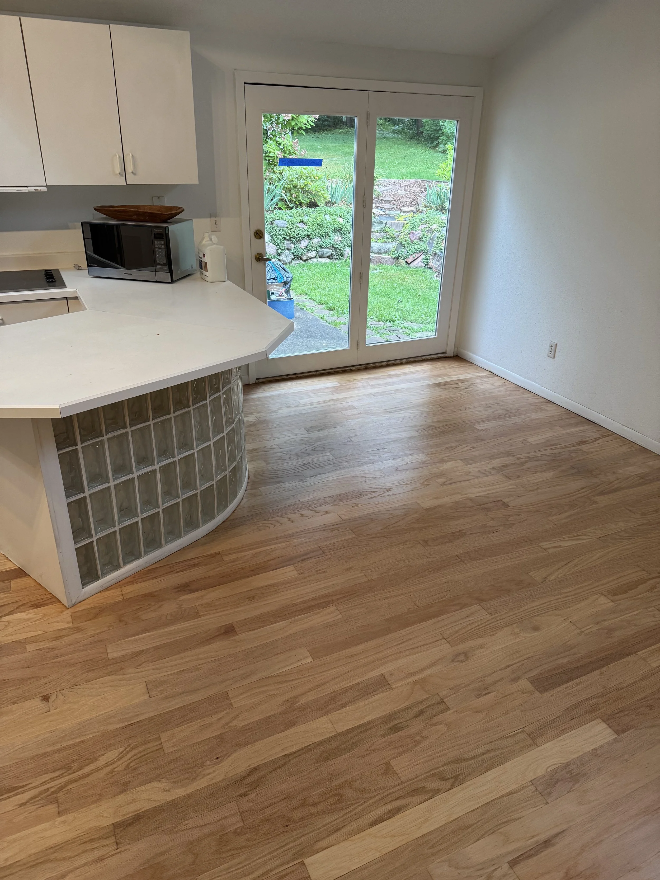 Empty kitchen area with wooden flooring, white cabinets, a microwave, and a glass block panel on the counter. Large glass sliding door leading to a backyard with grass and shrubbery.