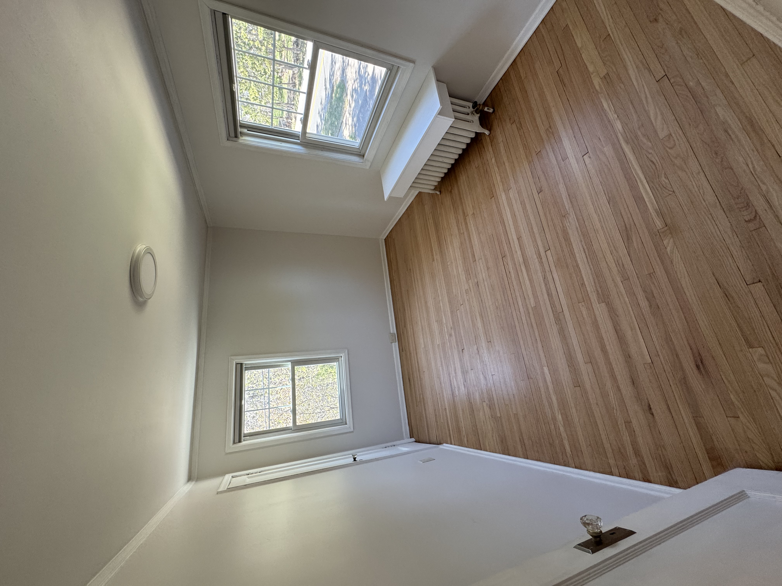 An empty room with wooden flooring, two windows showing outdoor trees, and a radiator beneath one window. The walls are painted white.