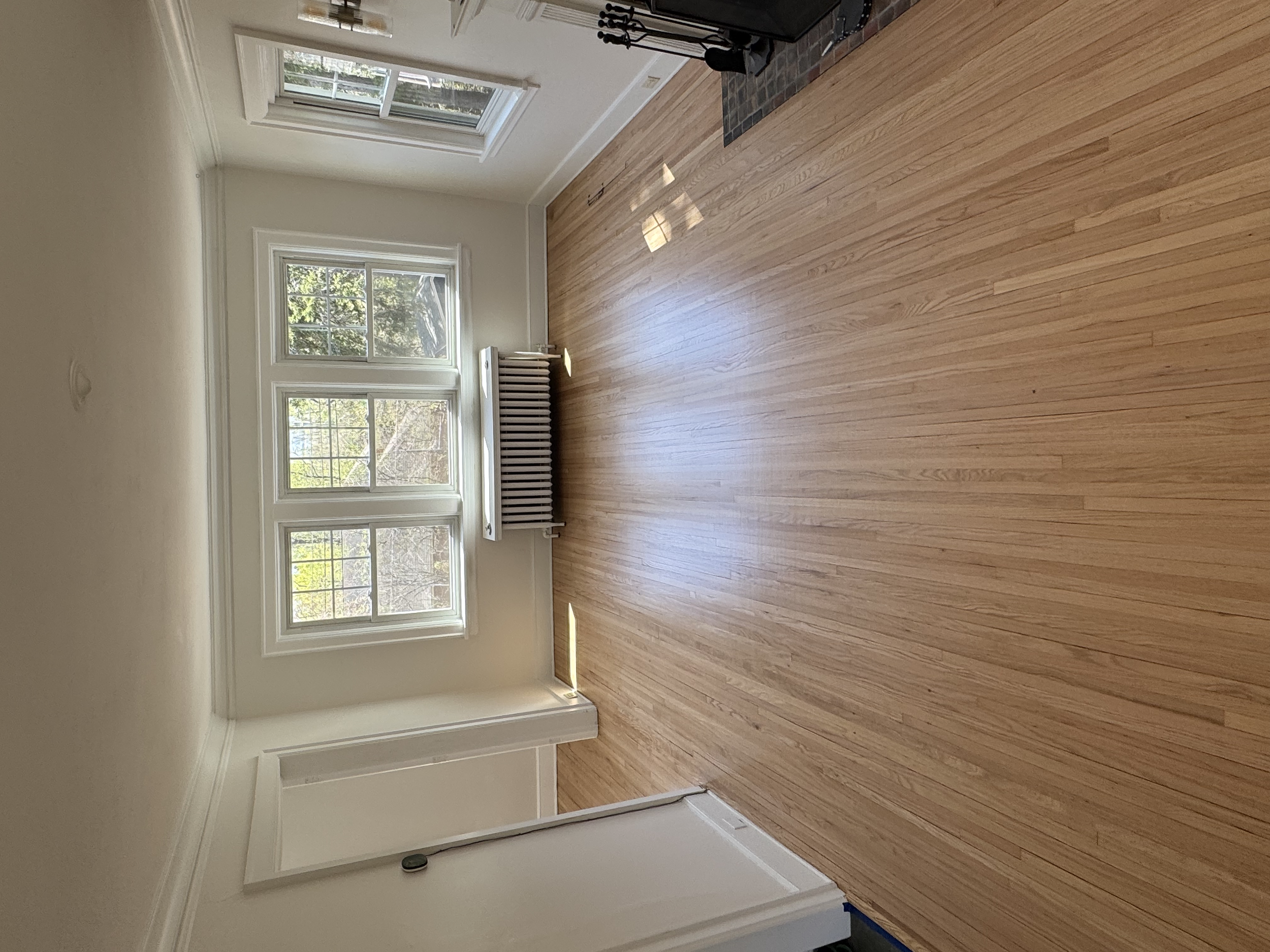 Empty living room with three large windows, a radiator beneath the central window, hardwood floors, and a fireplace with a black grate.