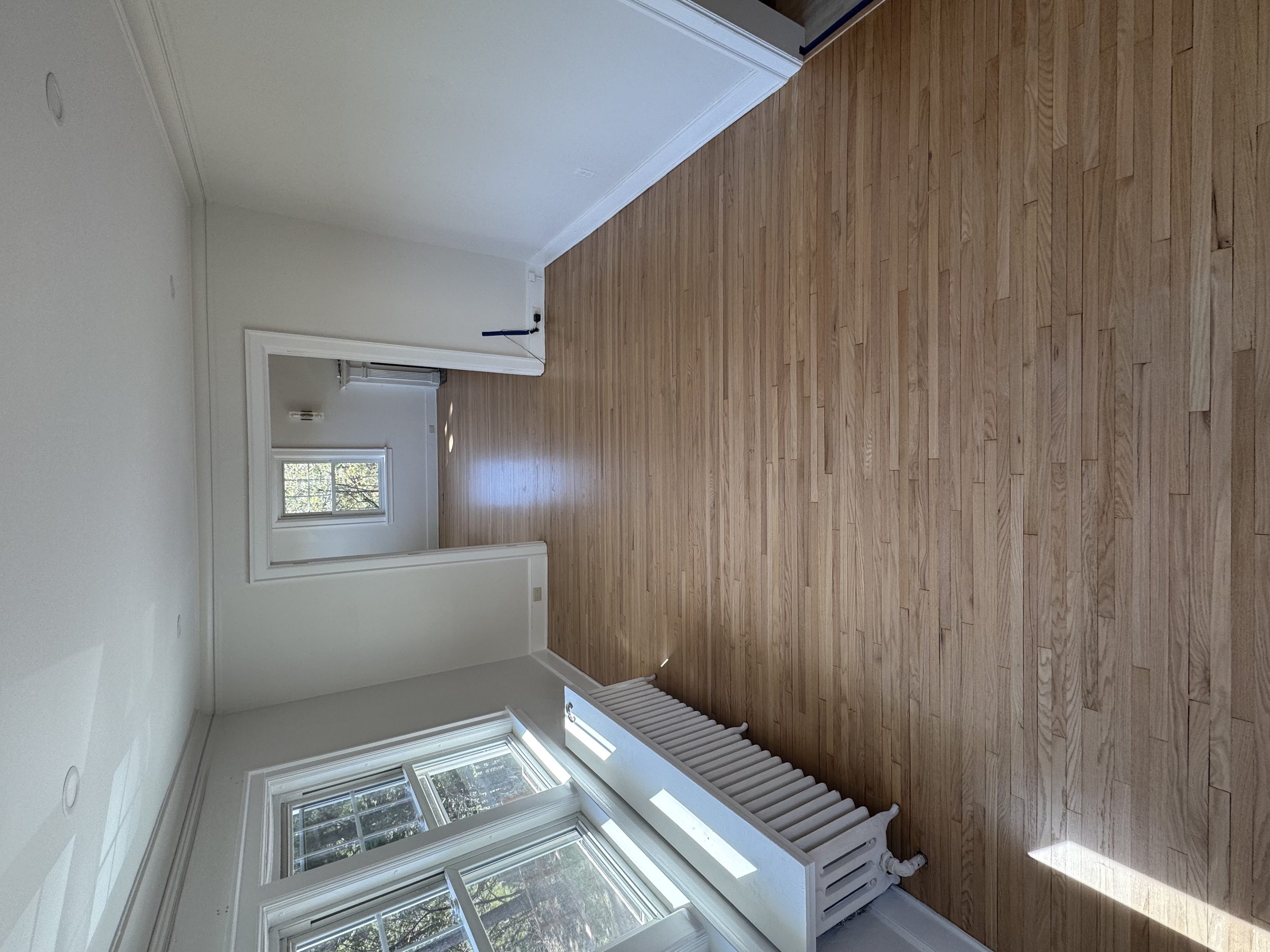 Empty room with hardwood floor, white walls, large windows, and a radiator.