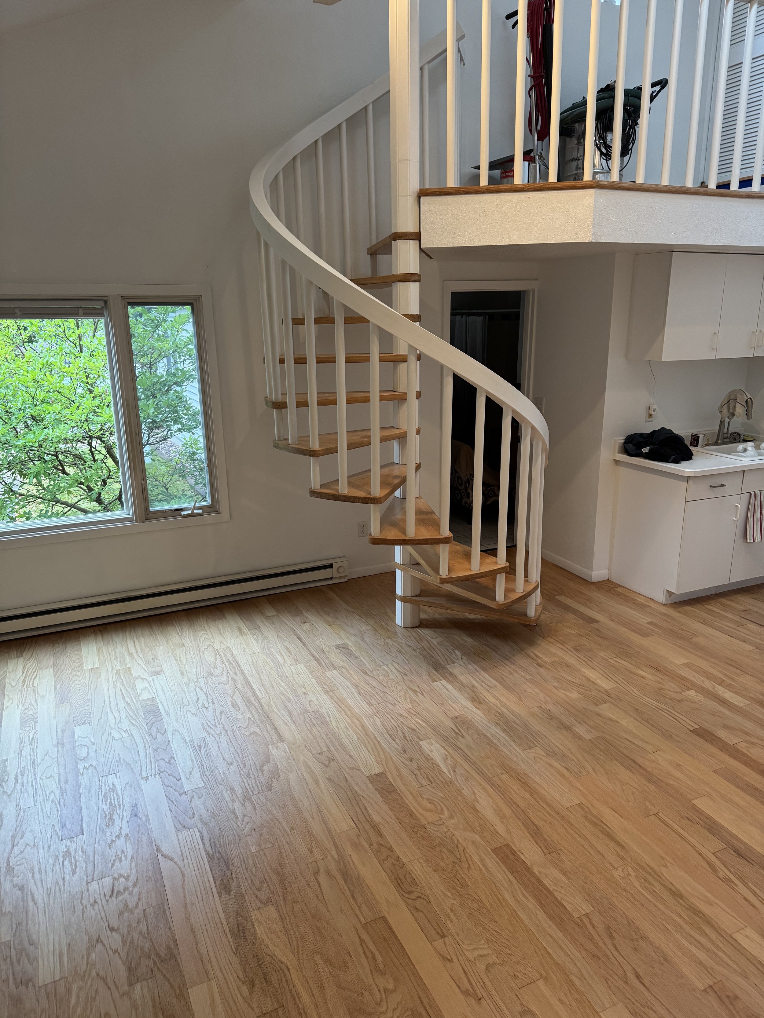 Room with hardwood flooring, a large window with greenery outside, a white spiral staircase leading to an upper level, and part of a kitchen with a white countertop and sink.
