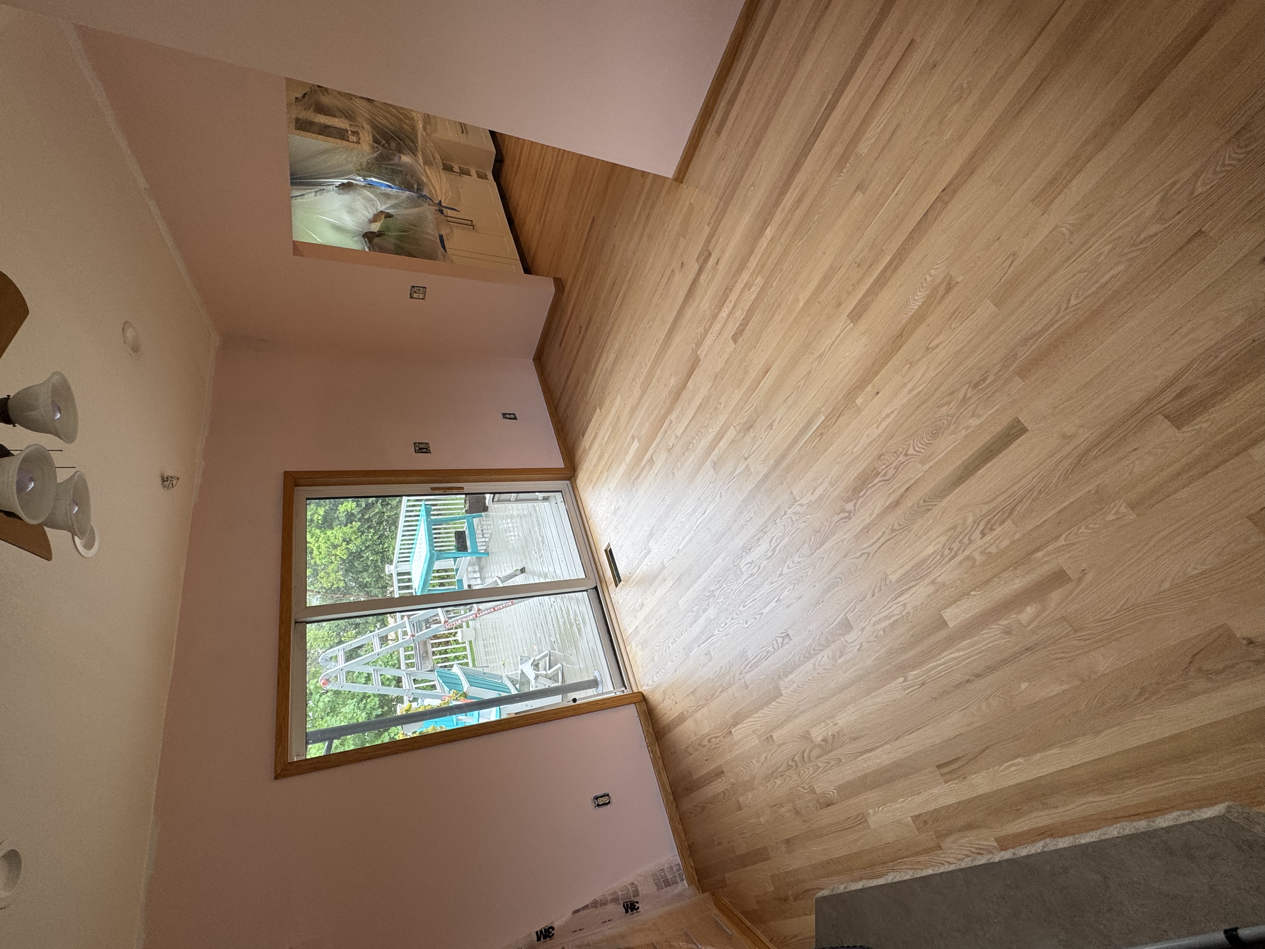 Empty living room with hardwood floors, sliding glass door leading to a deck with outdoor furniture and toys, and a glimpse into another room through an open wall.