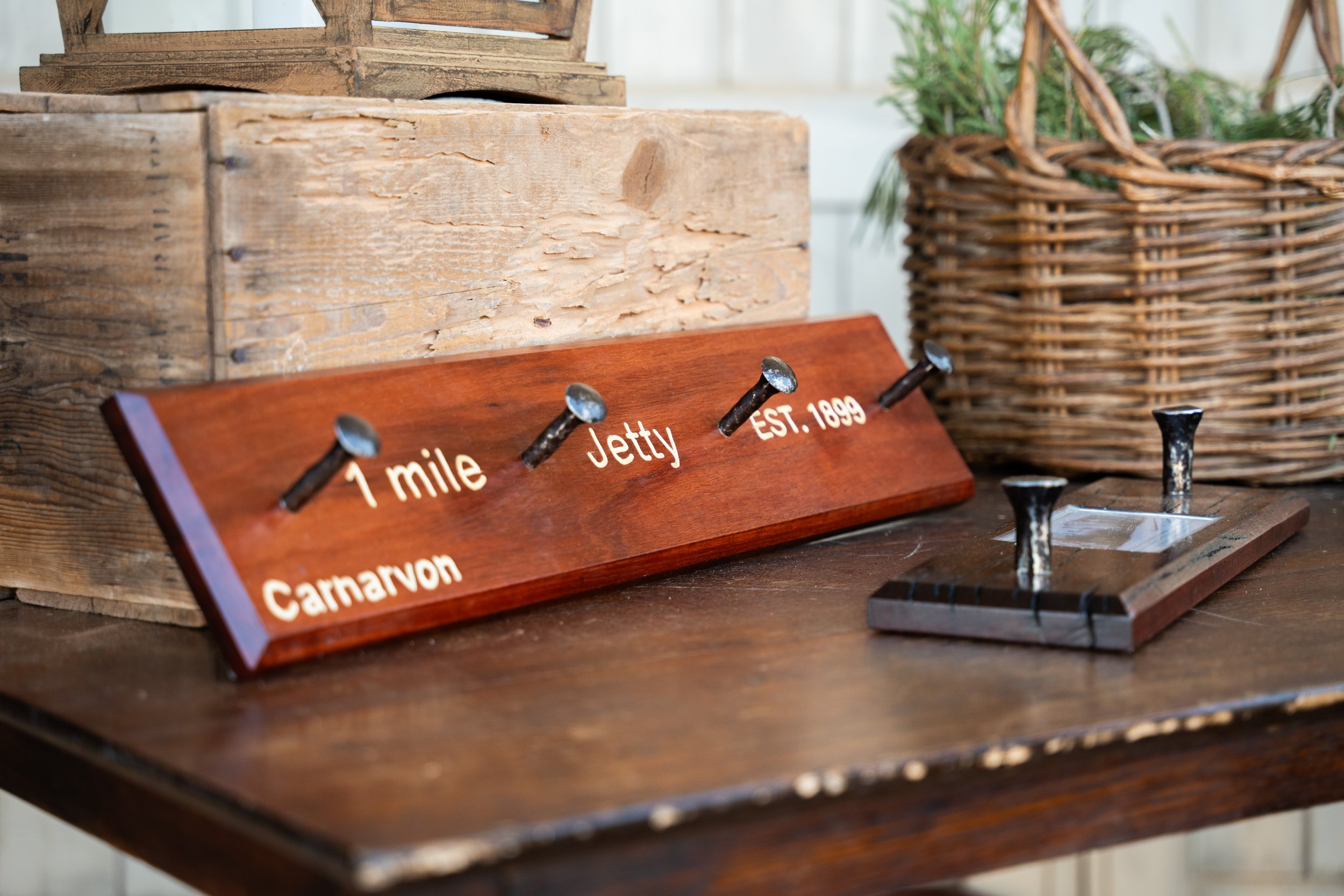 A decorative wooden sign with metal nails displaying the words '1 mile jetty,' with an additional nail pointing to 'Carnarvon' and 'EST. 1899,' placed on a wooden surface near a wicker basket.