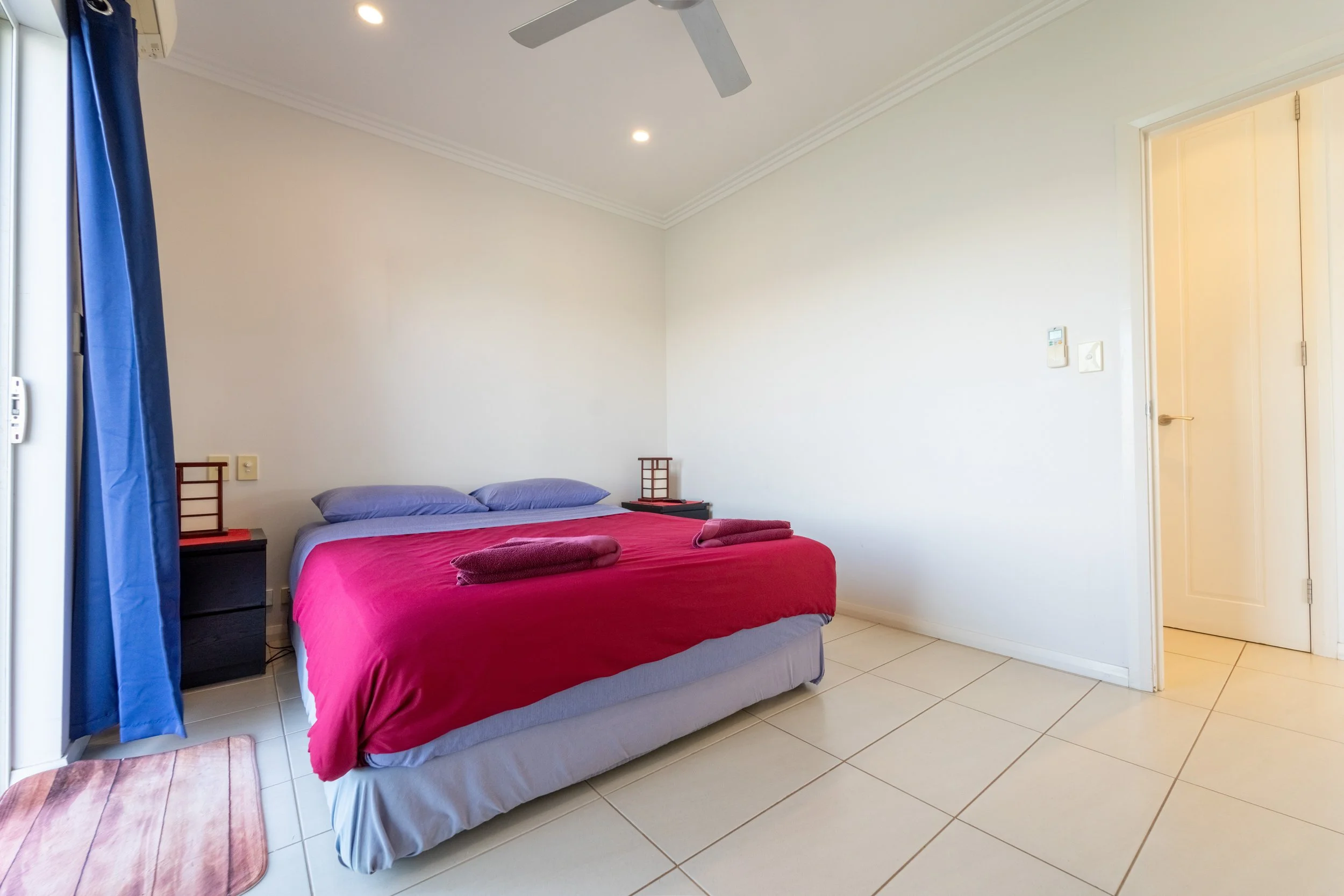A bedroom with a bed covered in red sheets and purple pillows, two folded towels on top, and a small nightstand on each side. Blue curtains are partially open, and there is a ceiling fan and recessed lighting. The floor has beige tiles.