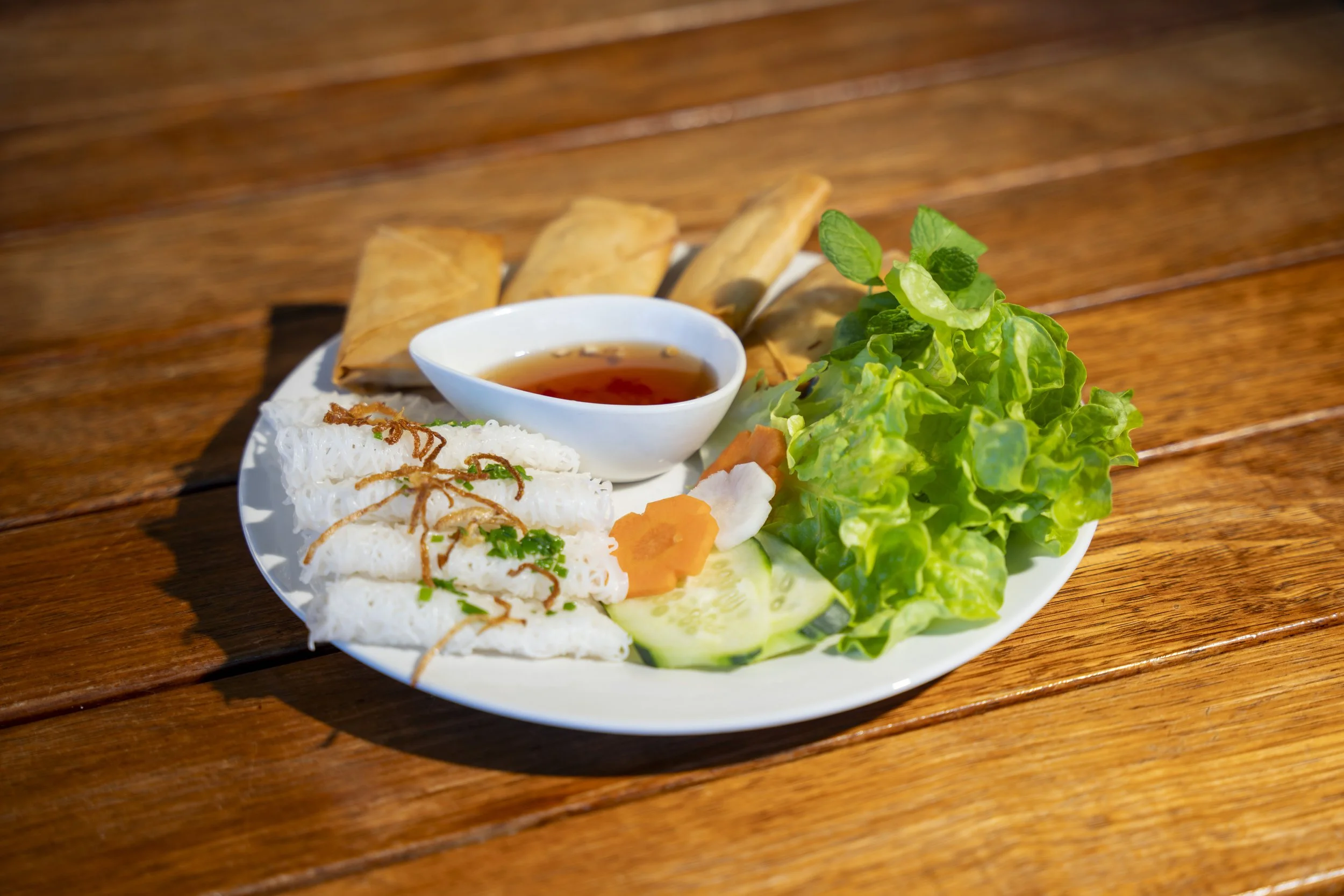 Vietnamese dish with rice paper rolls, fresh vegetables, fried egg rolls, and dipping sauce on a white plate on a wooden table.