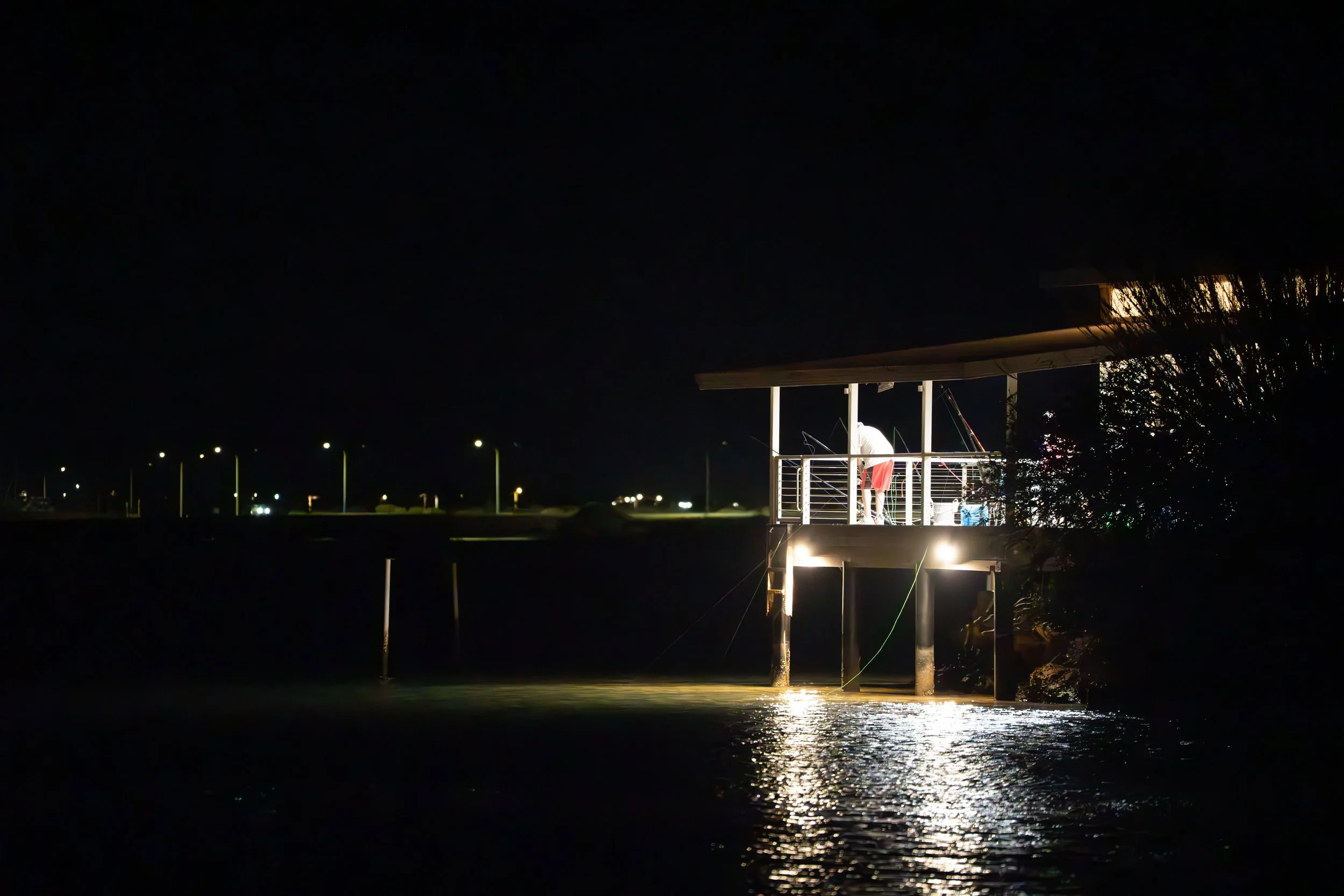 Nighttime scene of a small illuminated fishing pier or balcony with two people fishing, reflecting on calm water, with streetlights visible in the background.