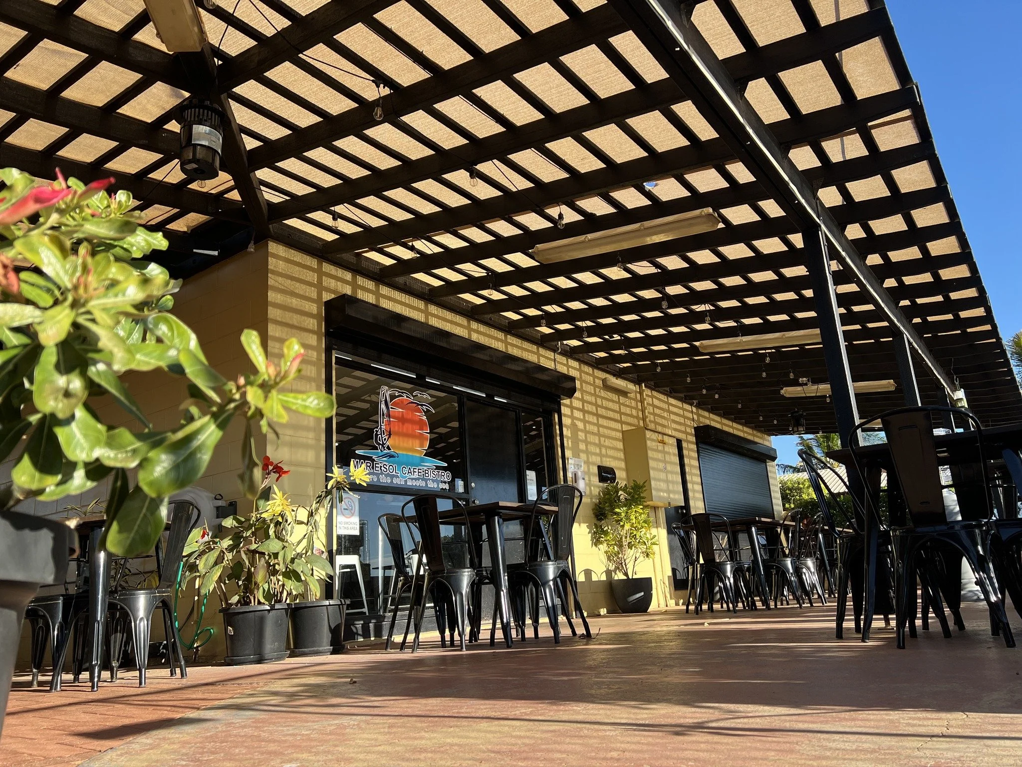 Outdoor seating area of a cafe or bistro with black chairs and tables, potted plants, and a shaded pergola with shadows cast on the wall during daytime.