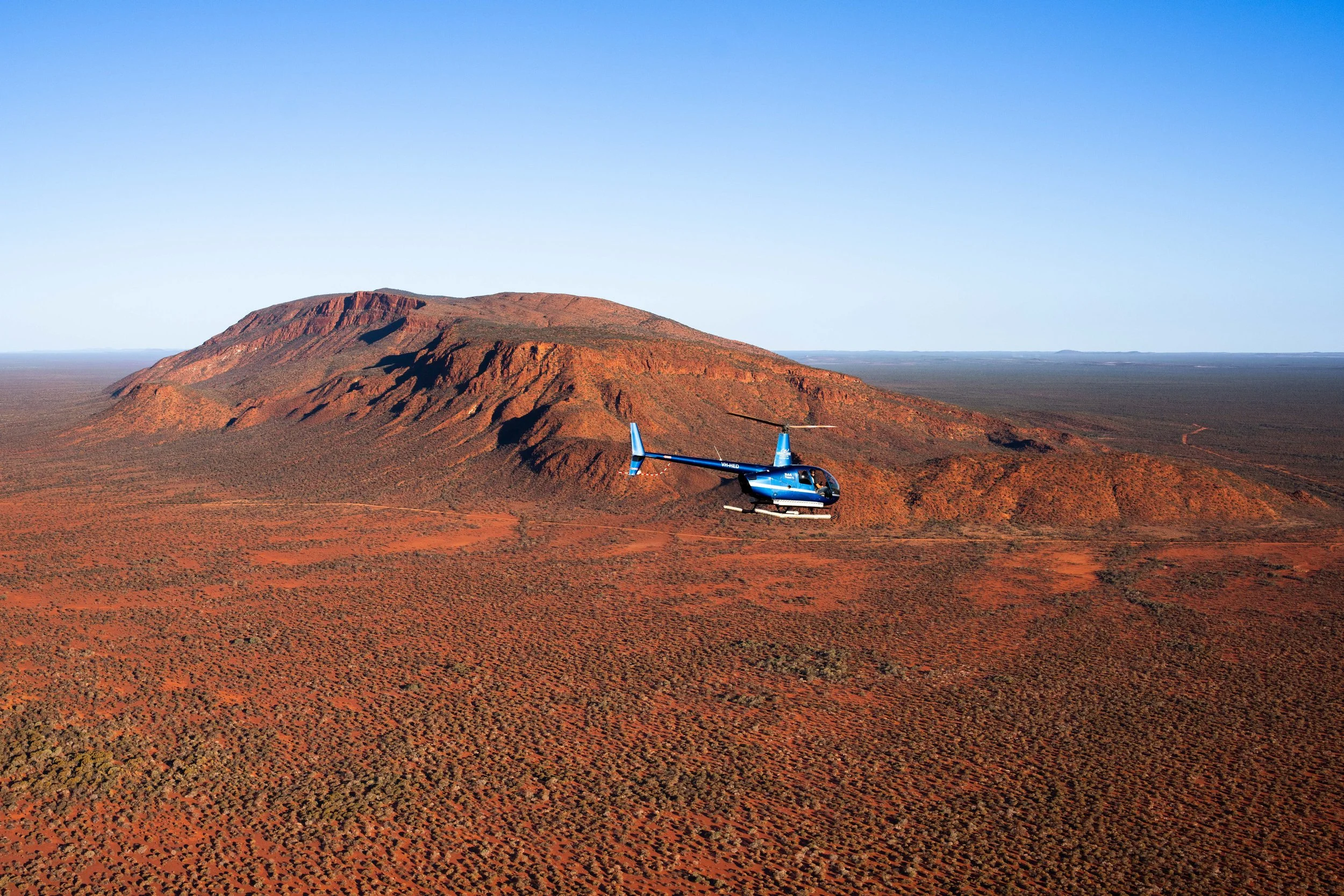 A blue helicopter flying over a red desert landscape with a large, flat-topped mountain in the background.