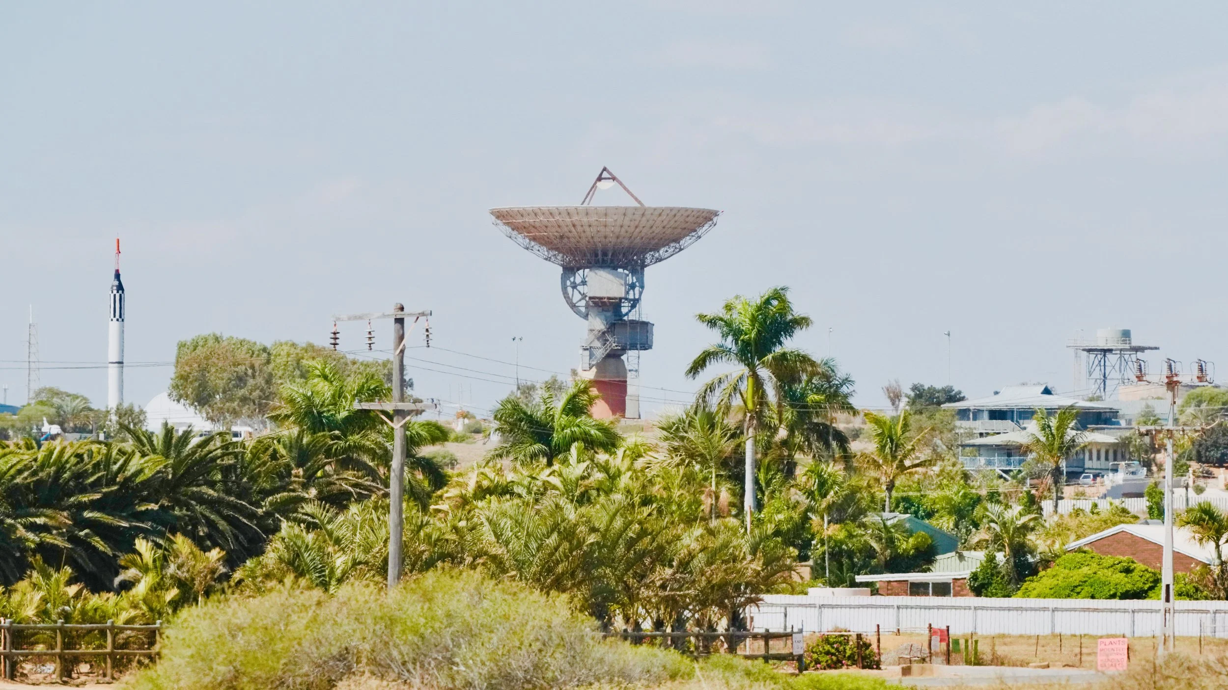 A landscape scene with lush green trees and palm trees in front of houses and buildings. In the background, there is a large satellite dish and communication towers, with a partly cloudy sky above.