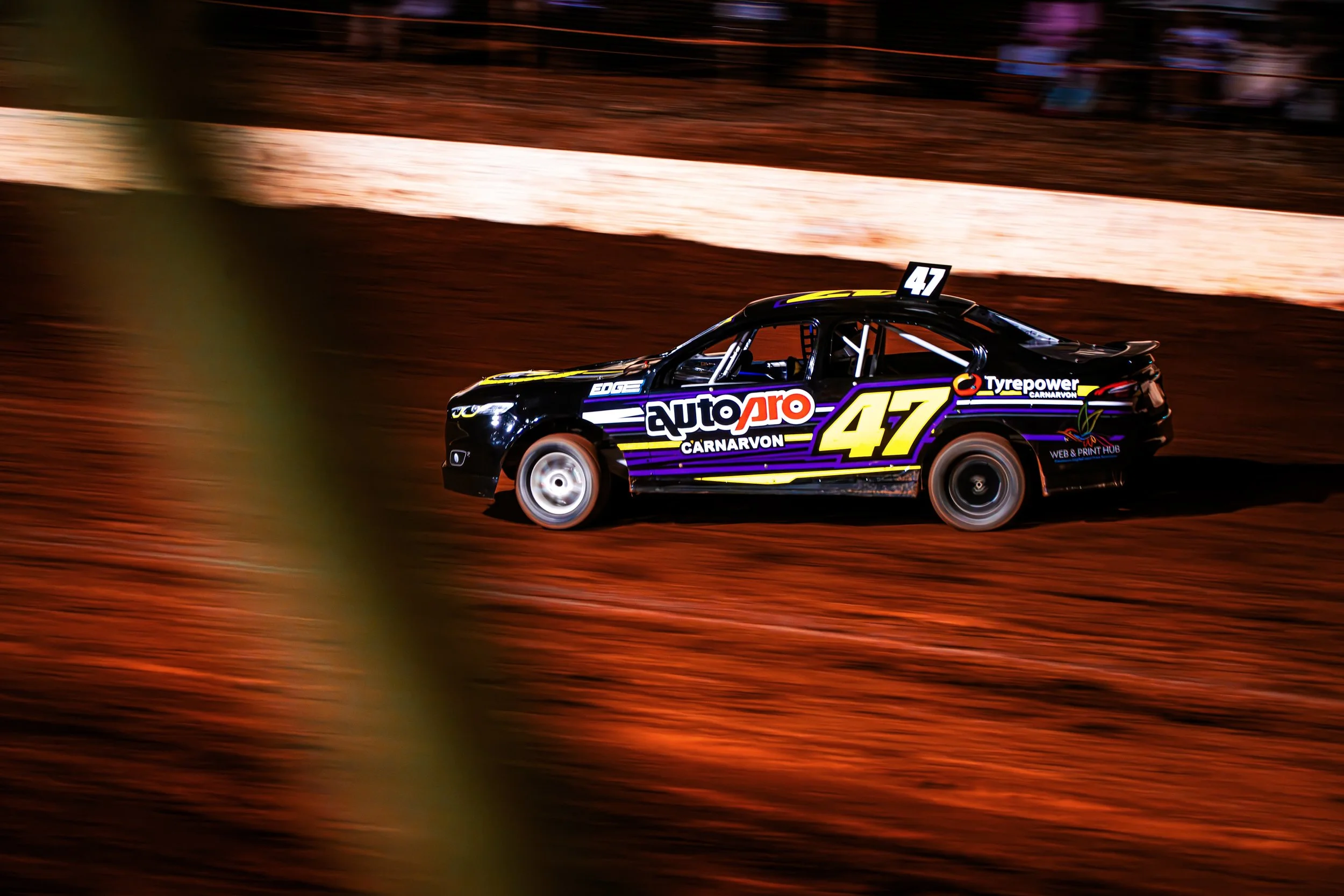 A black racing car with purple and yellow accents, number 47, speeding on a dirt track at night.