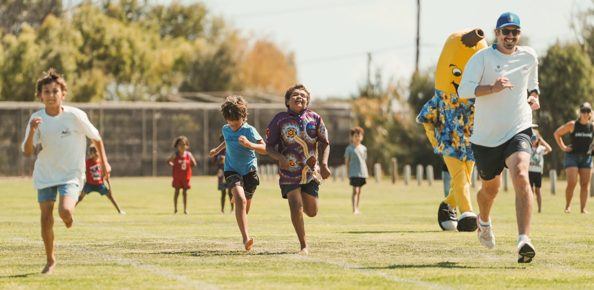 Children and adults running on a grassy field during an outdoor event, with some passing by a person in a banana costume in the background.