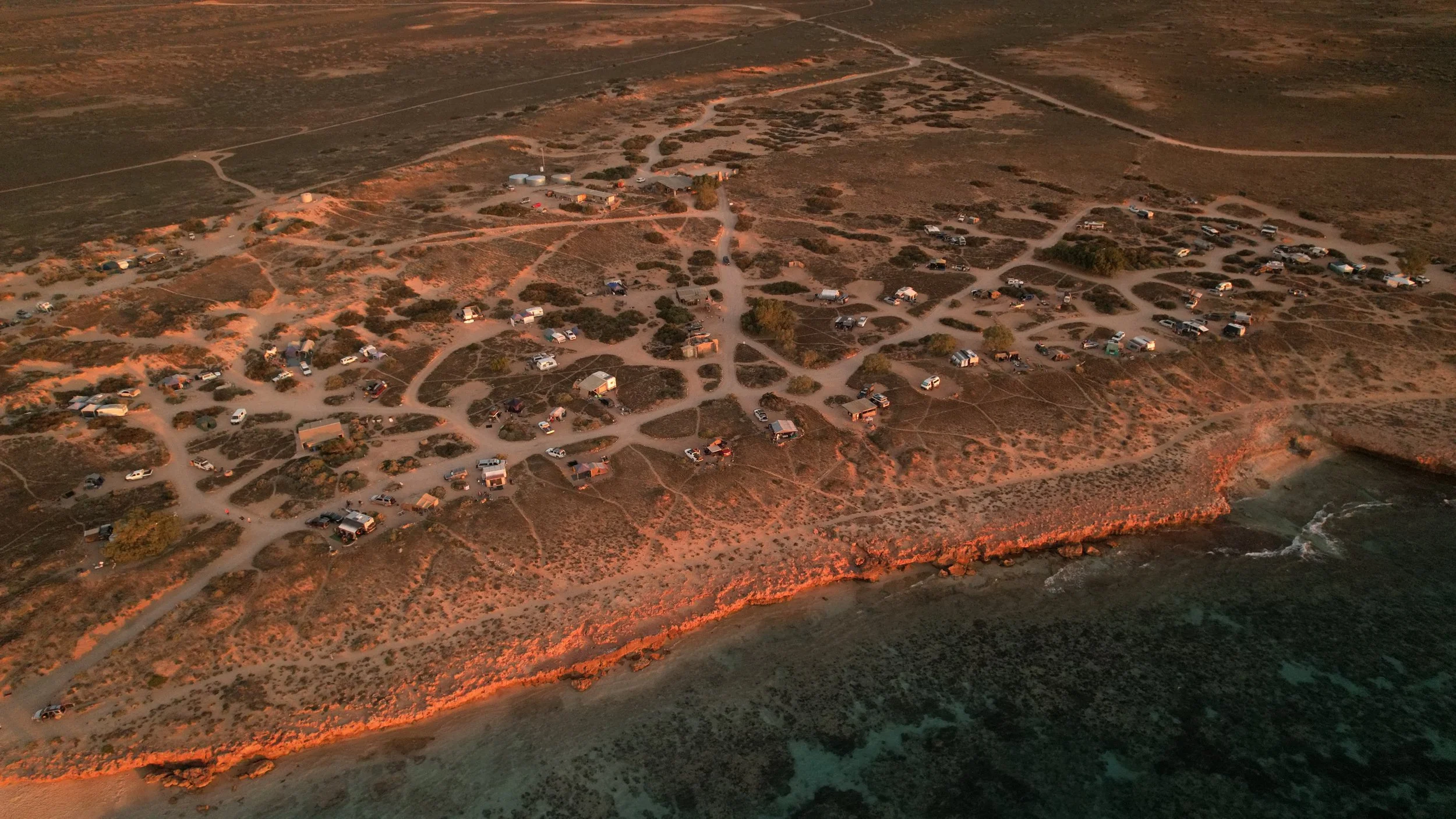 Aerial view of a coastal campground at sunset. The area has dirt roads, small trailers, tents, and vehicles scattered along the rocky shoreline with waves crashing on the coast.