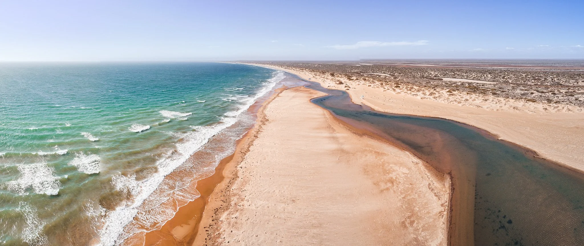 A beach with a shoreline on the left and a river flowing into the ocean on the right, under a clear blue sky.