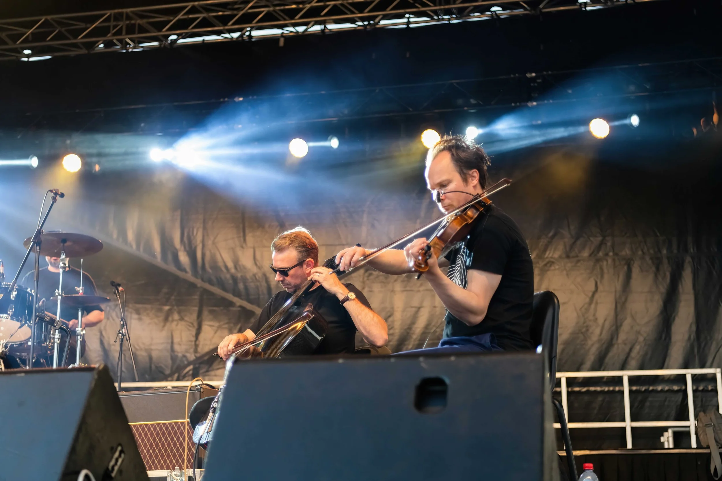Two musicians playing violin on stage during a live performance, with a drum set and stage lights in the background.