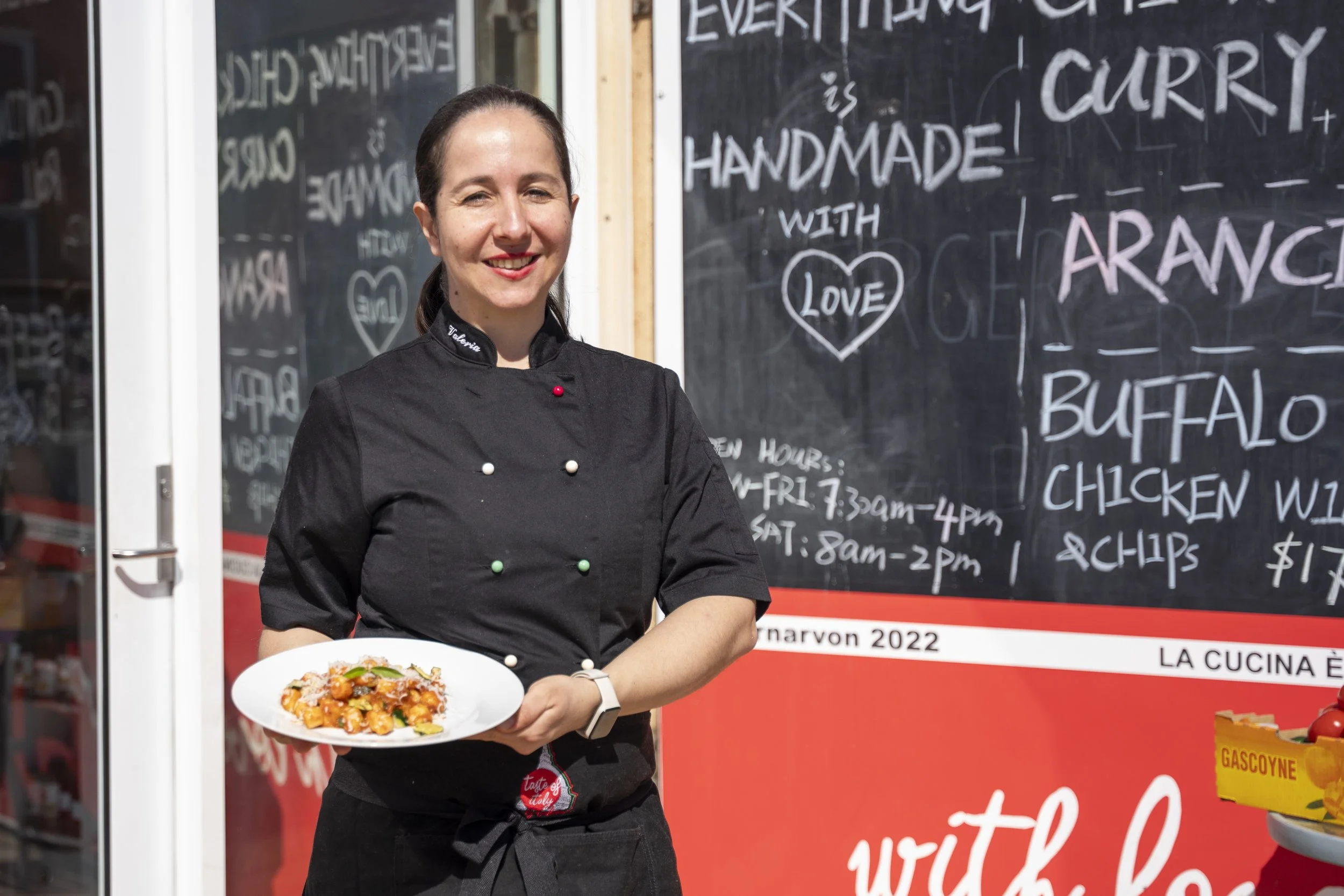 A smiling woman in a black chef's coat holding a white plate of pasta, standing outside a restaurant with a blackboard menu behind her.