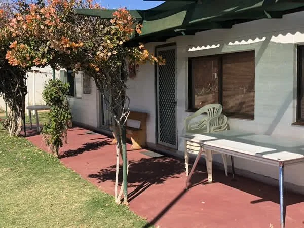 A backyard patio with a red concrete floor, a tree with pink flowers, a white plastic chair, a white table, and a house with a window and a door.