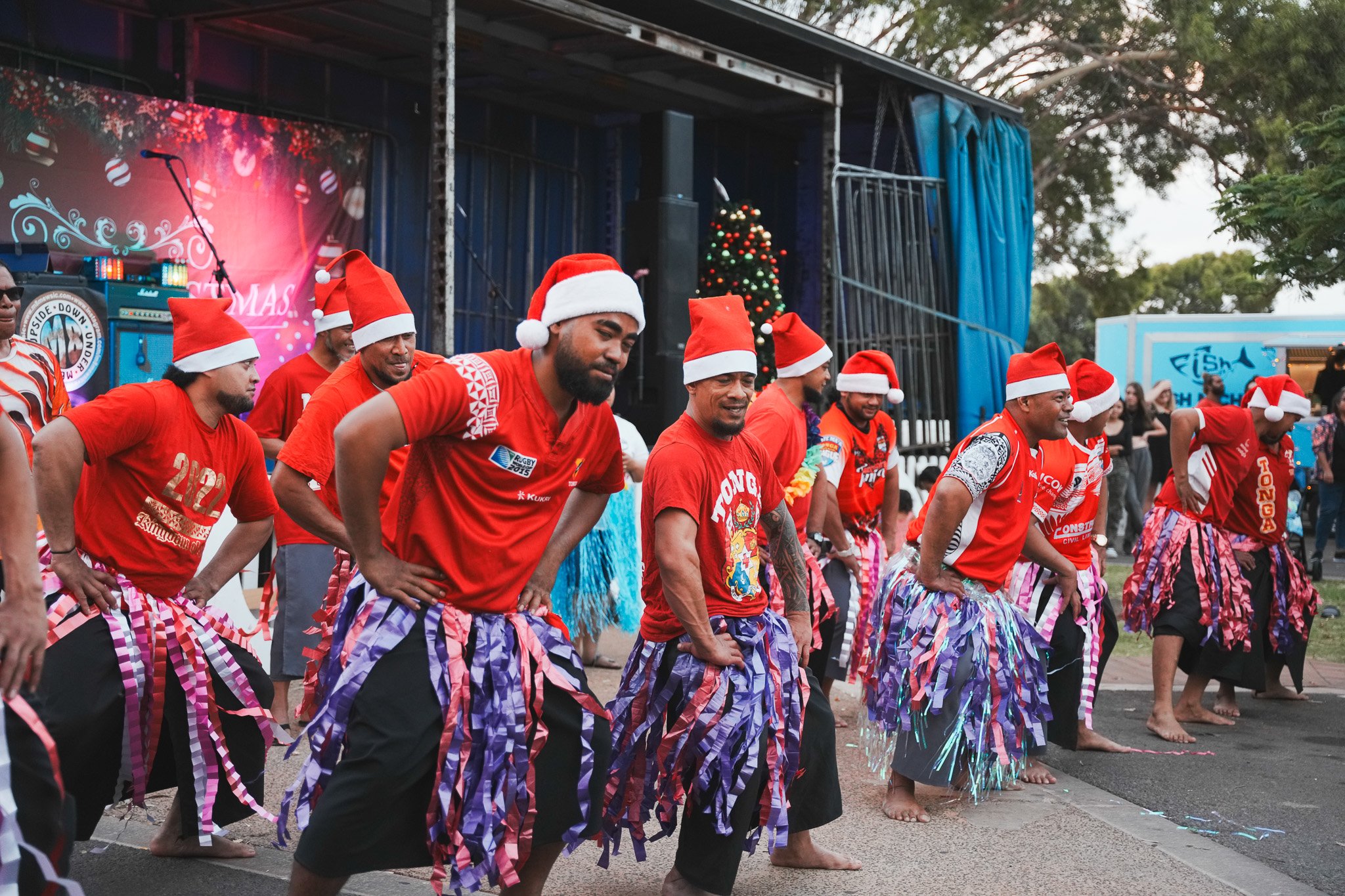 Group of men dancing in a Christmas-themed event, wearing Santa hats, red shirts, and colorful skirts, outdoors near a stage with Christmas decorations and a Christmas tree.