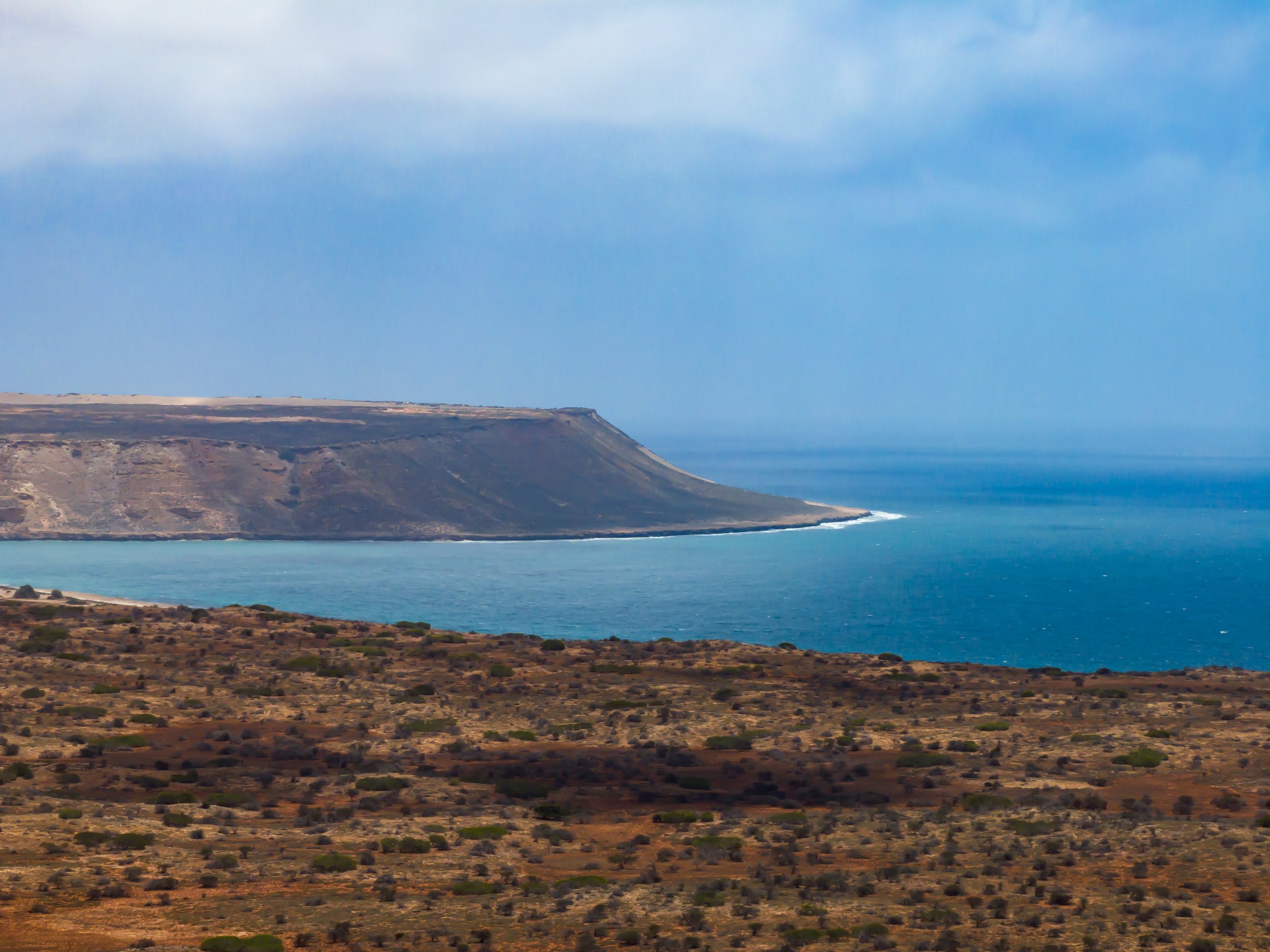 Landscape of a mountainous coastline with a large flat-topped cliff by the ocean, with dry, sparse vegetation in the foreground under a partly cloudy sky.