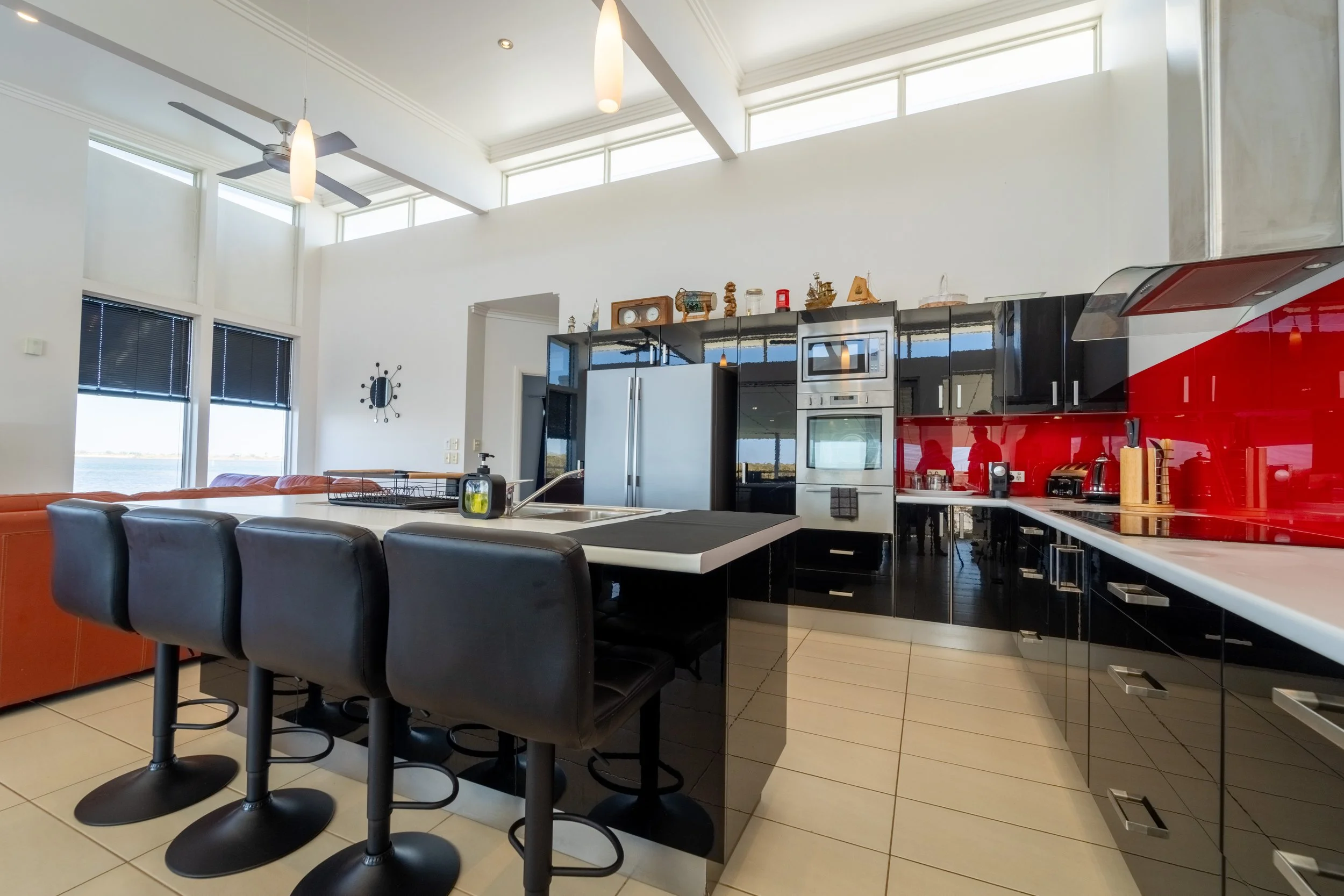 Modern kitchen with black glossy cabinets, red backsplash, stainless steel appliances, an island with four black barstools, large windows, and decorative items on top of the cabinets.