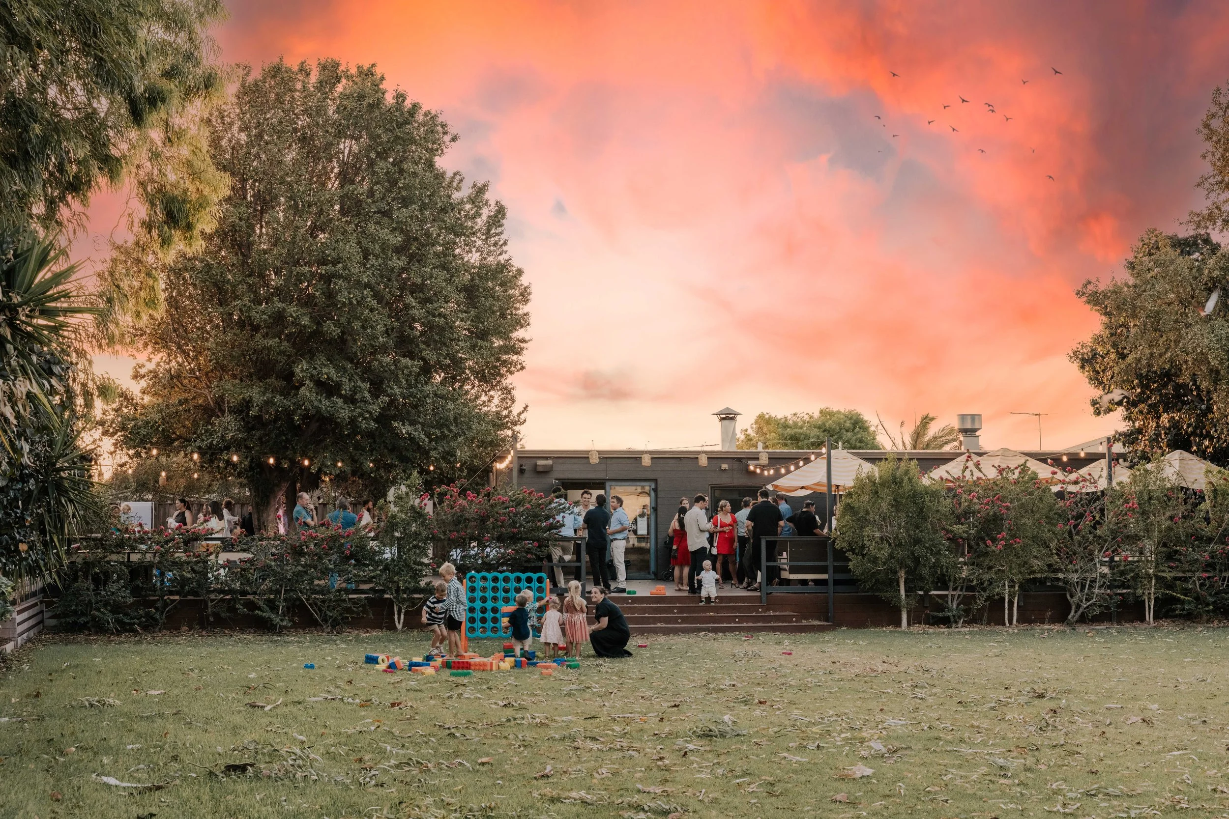 Outdoor gathering at a backyard patio during sunset with people socializing and children playing on the lawn.