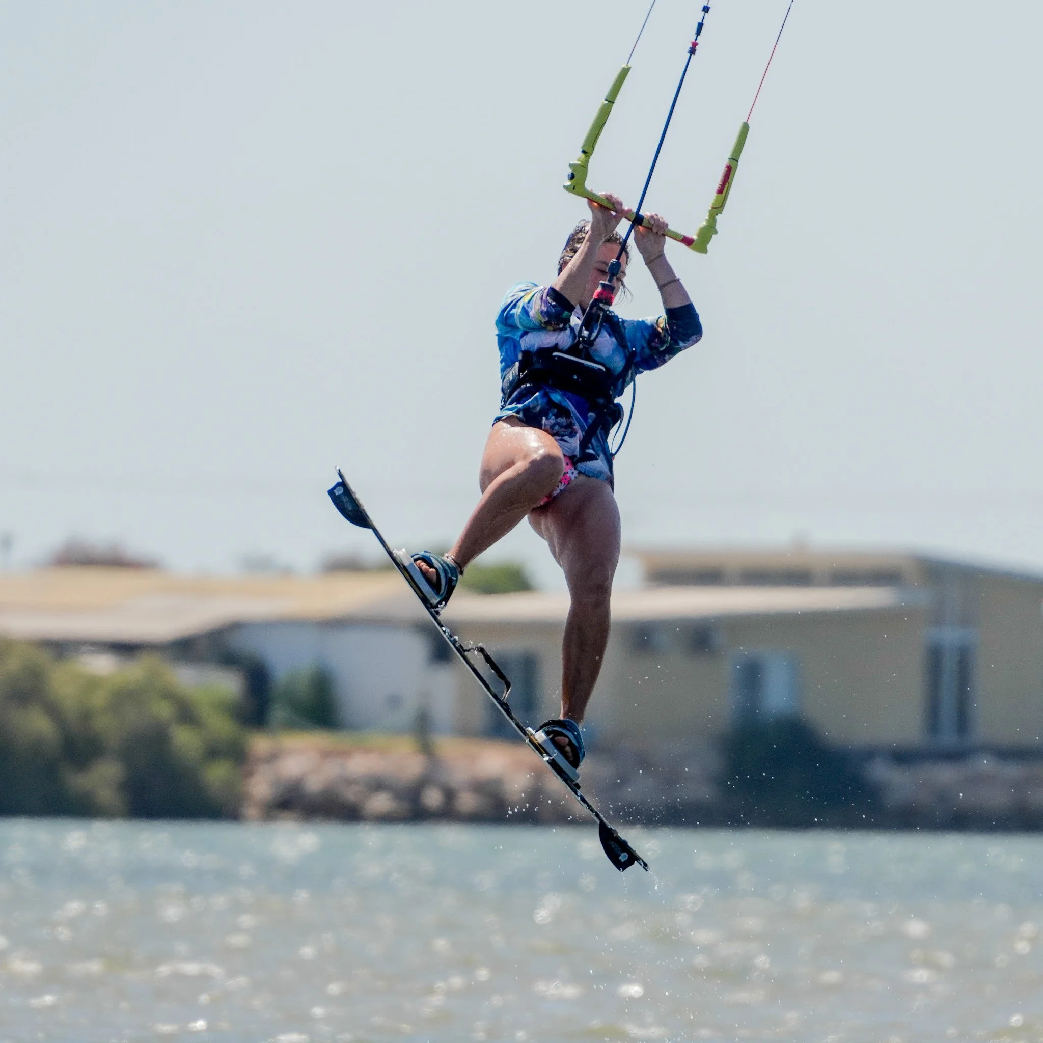 A person wakeskating on water while holding onto a kite, wearing a harness and colorful clothing, with houses and trees in the background.