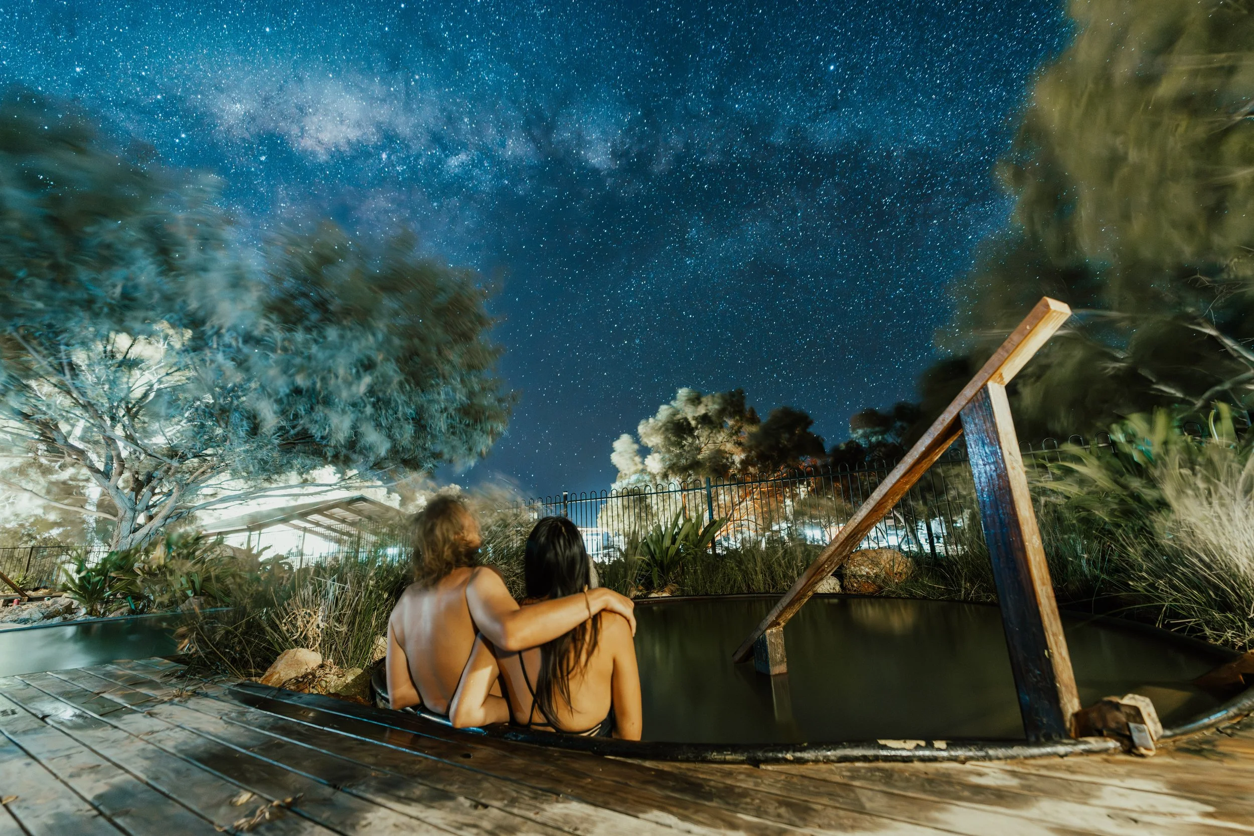 Two people sitting in a hot spring or pool at night, watching a star-filled sky with the Milky Way visible, surrounded by trees and foliage.