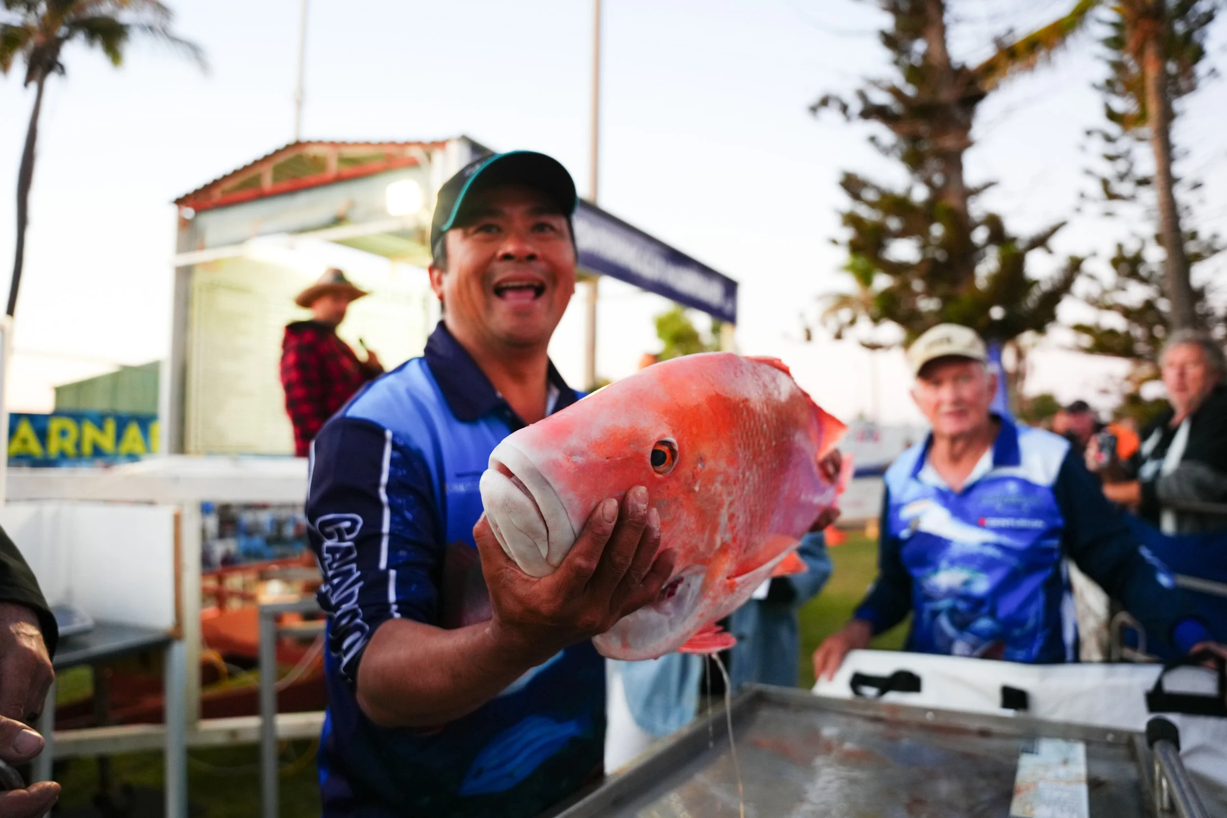 Man holding a large red fish at an outdoor event, with other people in the background.