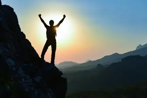 Person standing on a mountain cliff with arms raised at sunset.