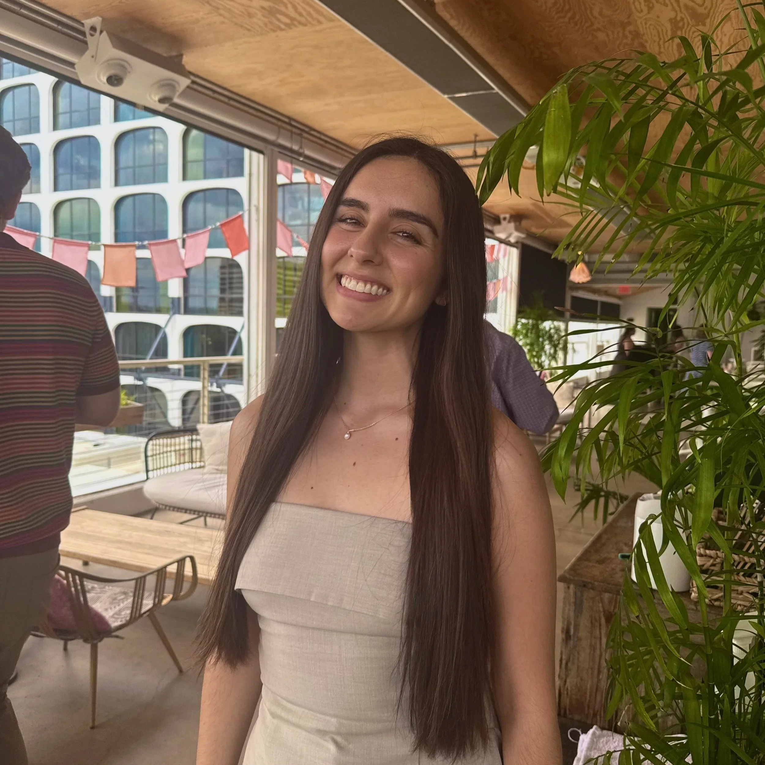 Smiling woman with long brown hair wearing a pearl necklace and strapless top, indoors with large windows, a green plant, and stringed flags in the background.