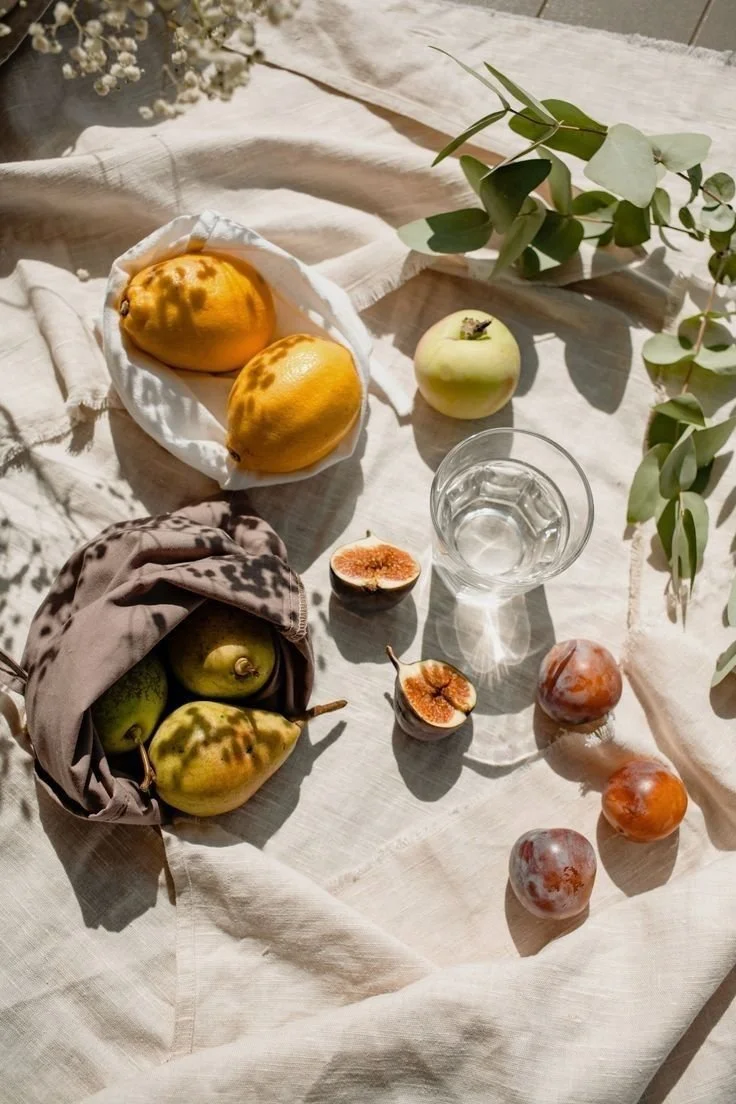 A still life scene with mixed fruits including pears, figs, an apple, and plums on a white cloth with green foliage.