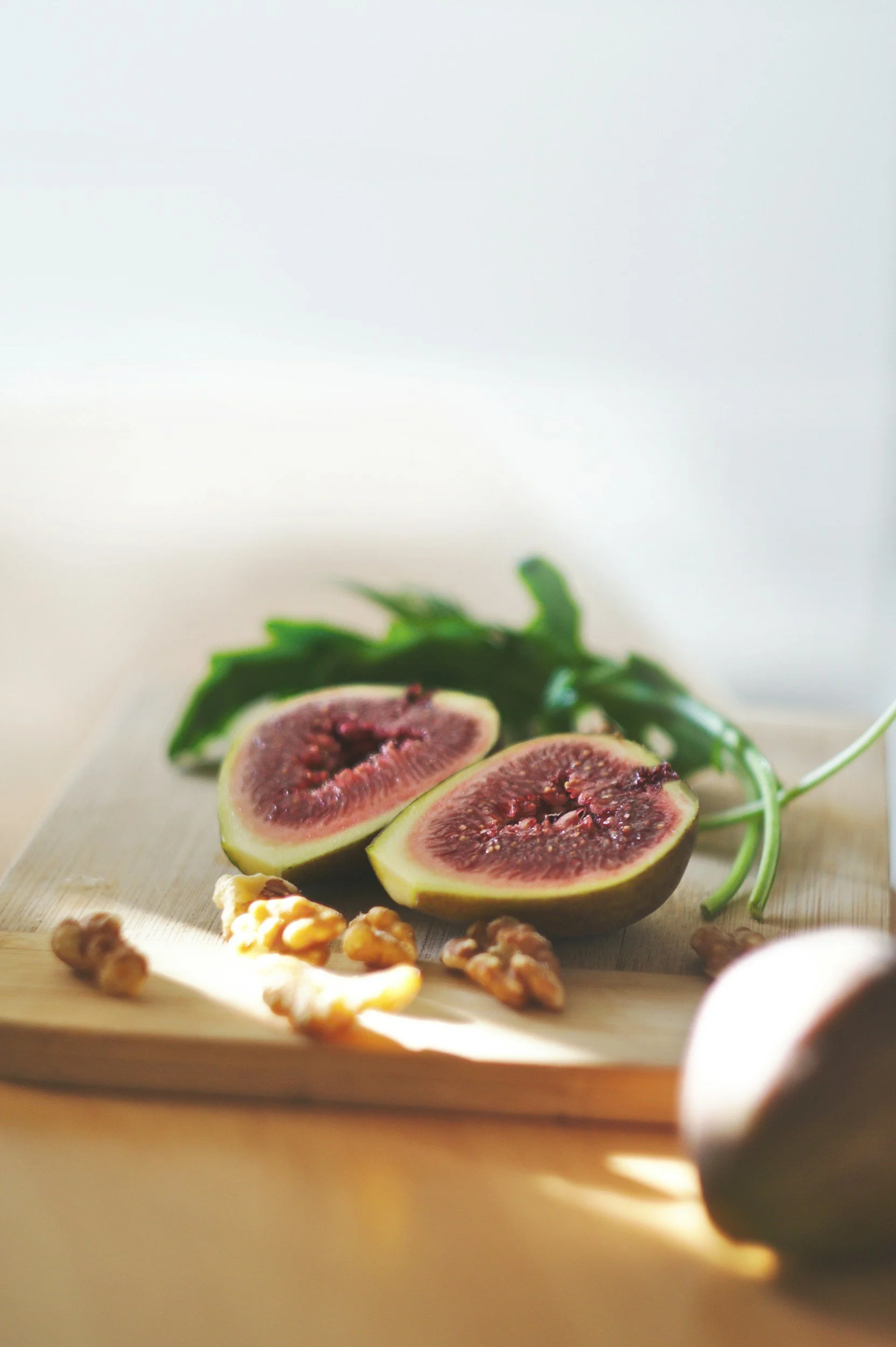 Close-up of a halved fresh fig with a dark purple exterior and pink interior, placed on a wooden cutting board with scattered walnuts and a few green leaves in the background.
