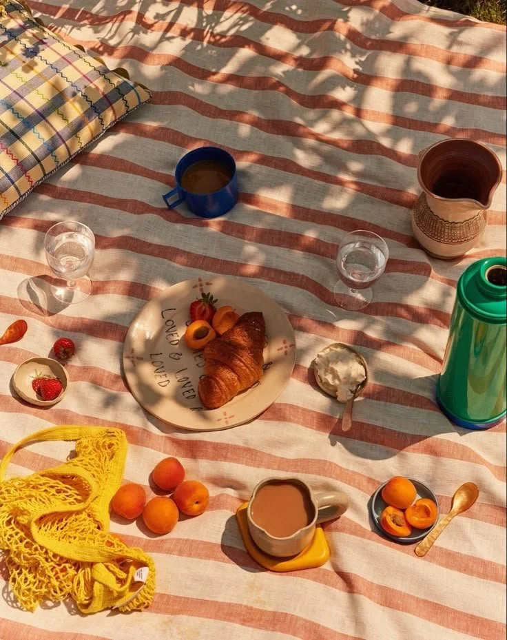 A picnic table with a striped tablecloth, a croissant with strawberries and apricots, a coffee cup, a small bowl of whipped cream, a yogurt container, a blue mug, a plastic green water bottle, glasses of water, a yellow net bag, a small plate with apricots, a spoon, and a decorative pitcher.
