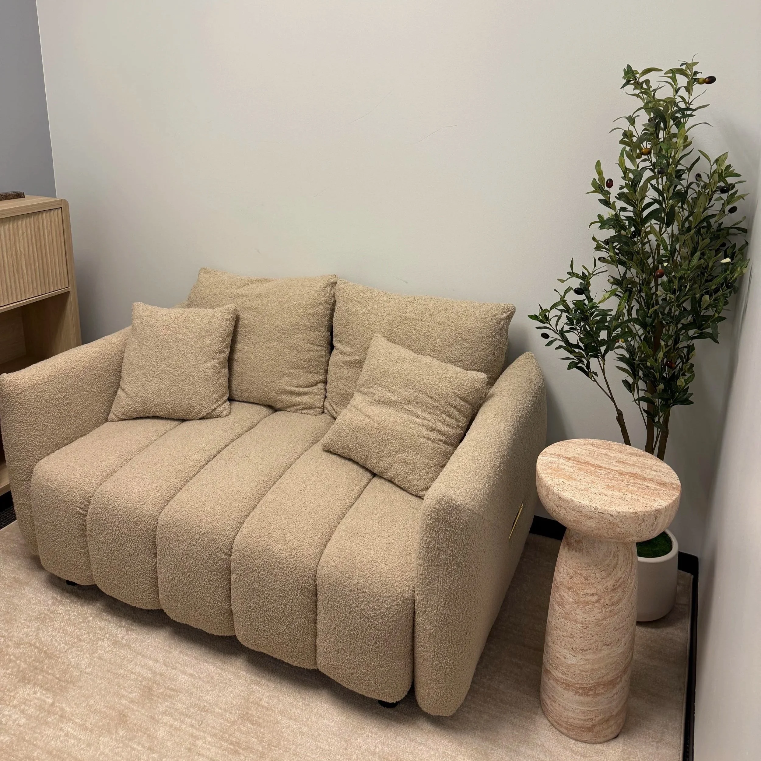 Beige sofa with multiple cushions, next to a side table with a rounded pedestal base, in a room with beige carpet and a white wall.