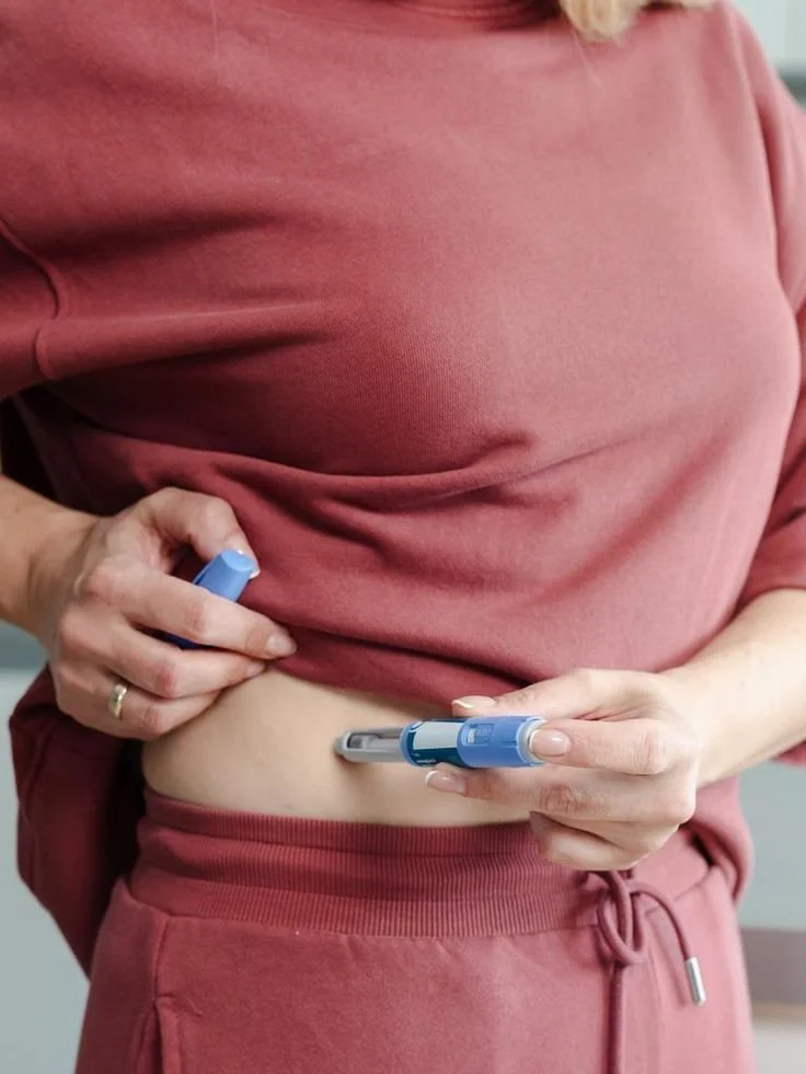 A woman wearing a pink shirt and matching pink pants holds a blue insulin pen and a transmitter for blood sugar testing, suggesting she is checking her blood sugar levels.