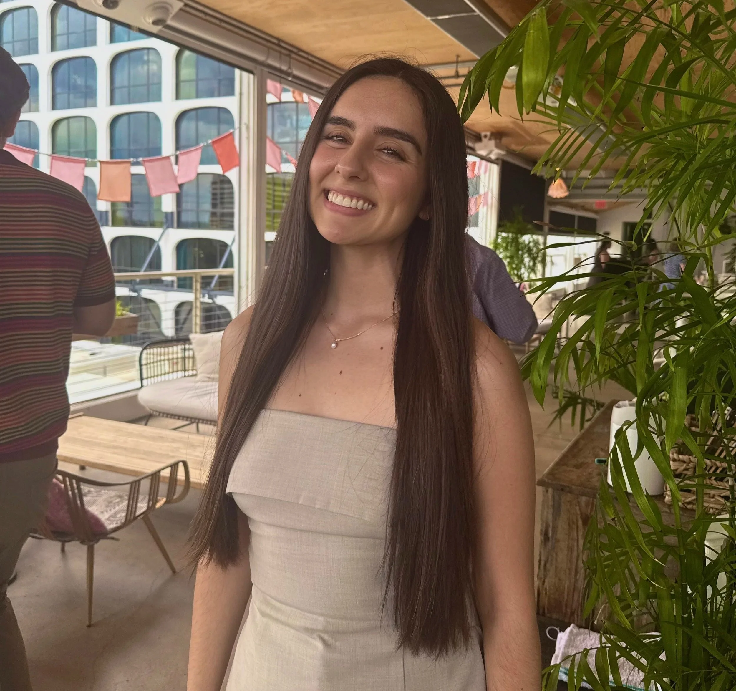 A woman with long dark hair smiling, wearing a light-colored sleeveless top and a necklace, standing indoors near green plants, with windows and modern buildings in the background.