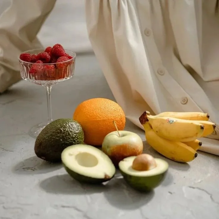 Assorted fruits including strawberries in a glass bowl, an orange, an avocado, green apple, banana bunch, and a halved avocado with a seed on a light-colored surface with a cream-colored shirt in the background.