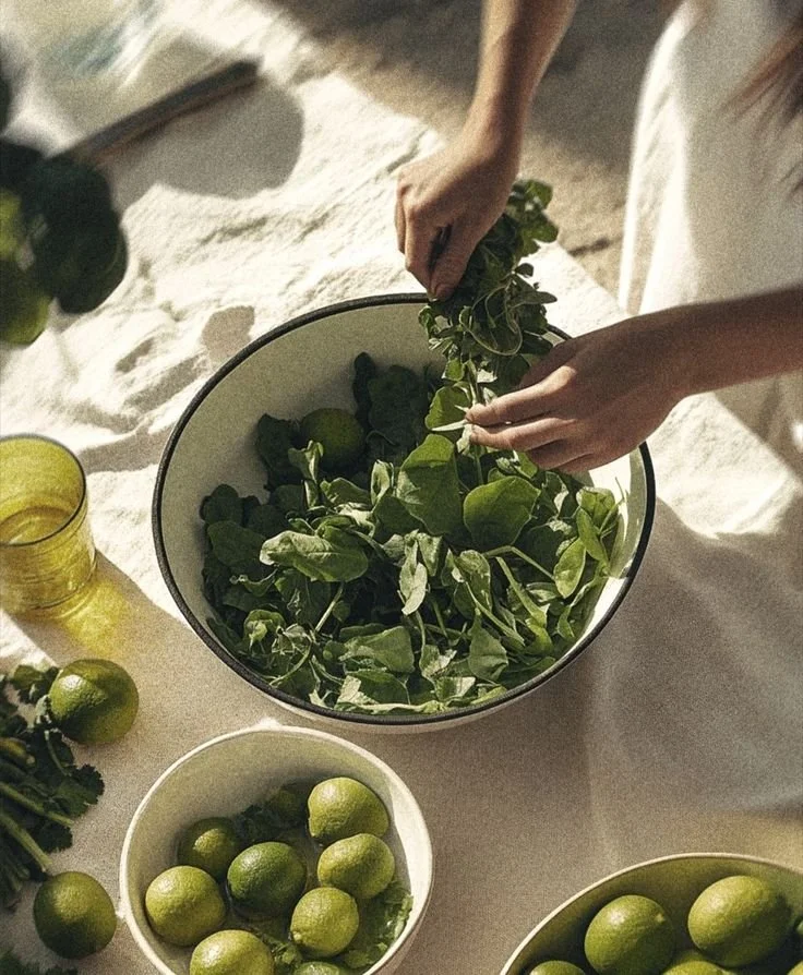A person preparing fresh herbs and lime for cooking or garnish on a white table, with bowls of limes and leafy greens.