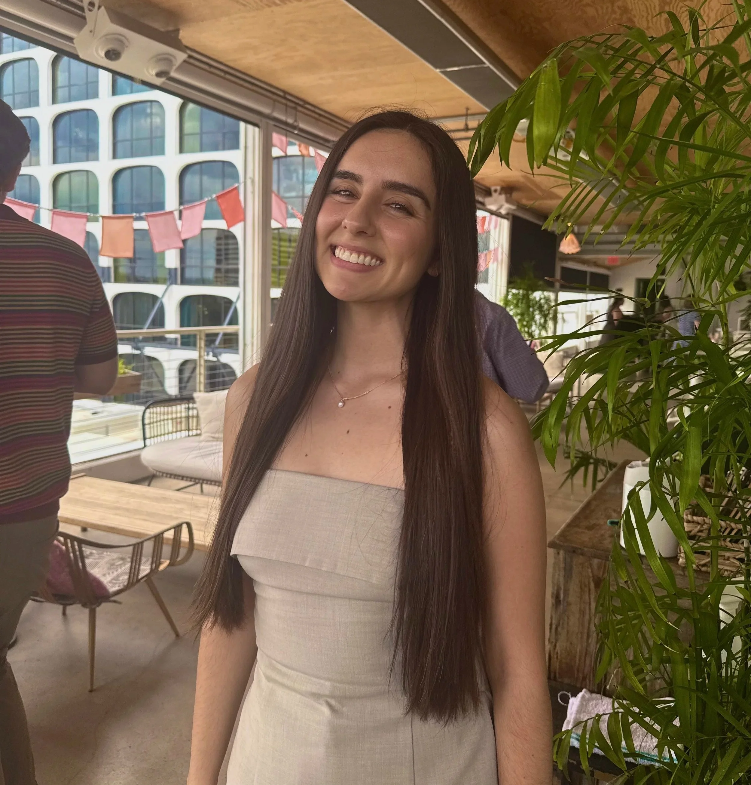 A woman with long dark hair, smiling, wearing a light-colored sleeveless top, sitting indoors near a large green plant, with a background of large windows and decorated red and orange flags.