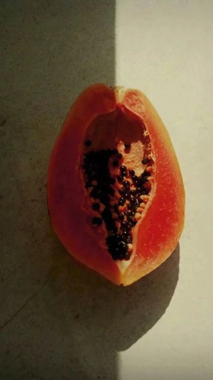 Half of a papaya with black seeds on a dark surface, casting a shadow on a light background.