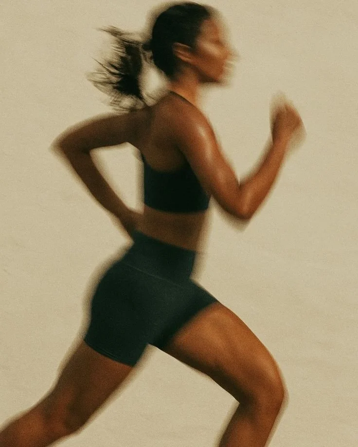A woman running, wearing a sports bra and shorts, with her hair tied back and veins visible on her arms.