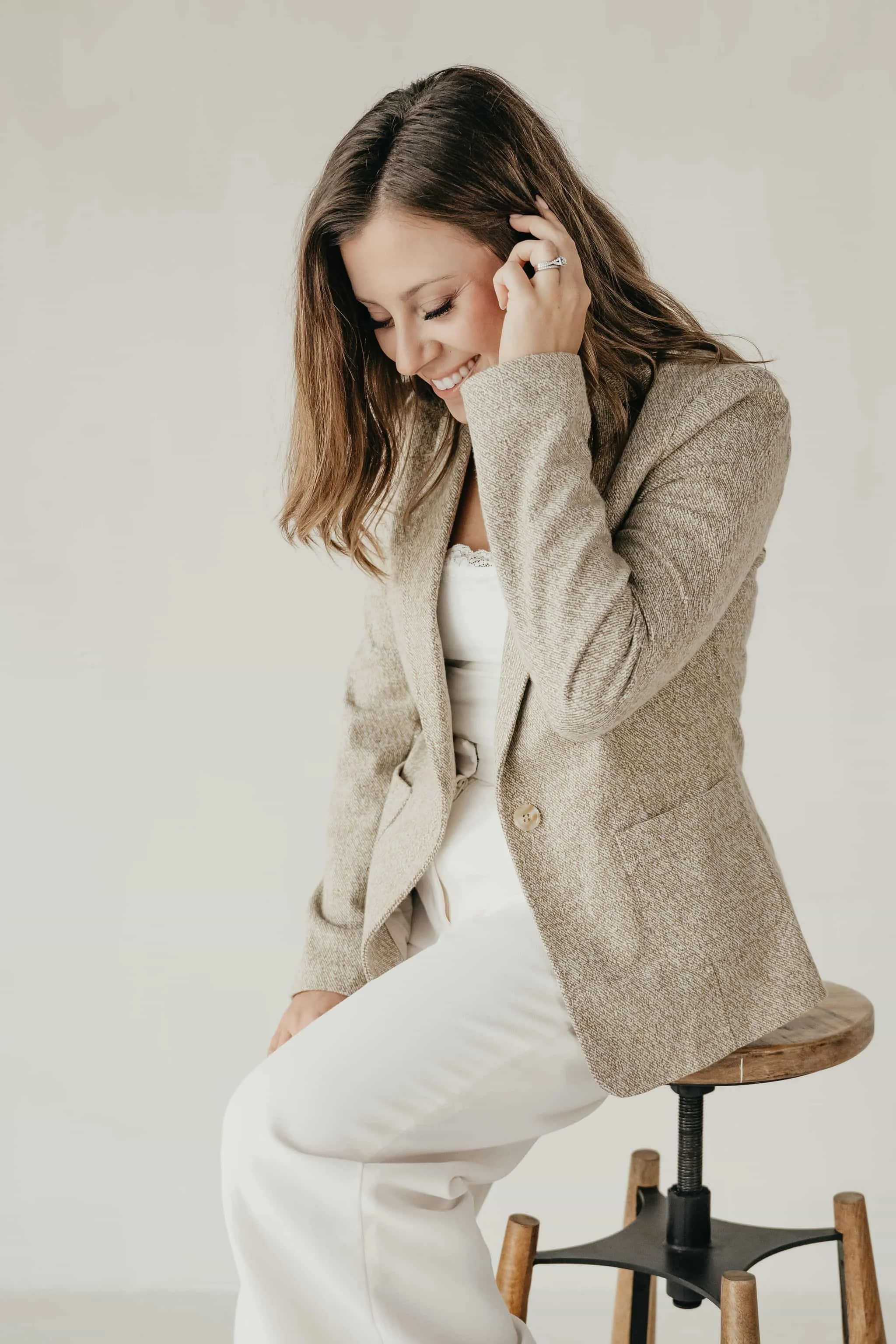 A woman with shoulder-length brown hair, smiling, wearing a beige blazer and white pants, sitting on a wooden stool against a plain light background.