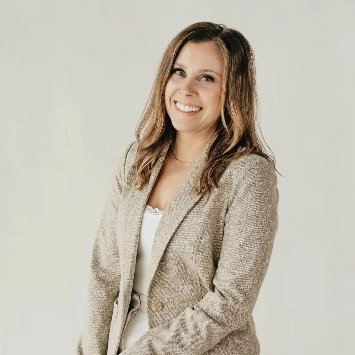 Portrait of a smiling woman with shoulder-length brown hair, dressed in a beige blazer over a white top, standing against a plain light background.