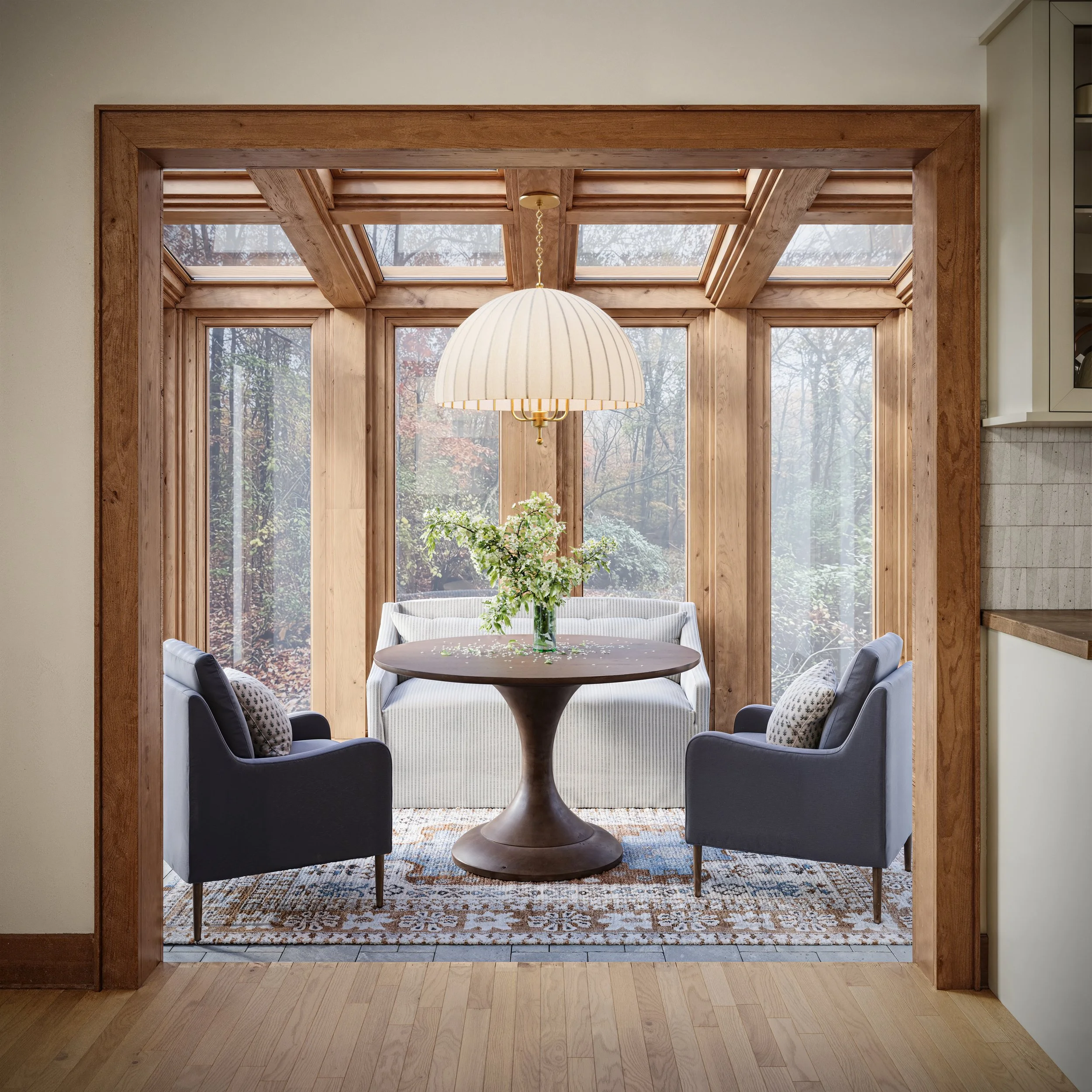 Cozy sunroom with large wooden framed windows, a round table with a floral centerpiece, and blue chairs, with a pendant light hanging above.