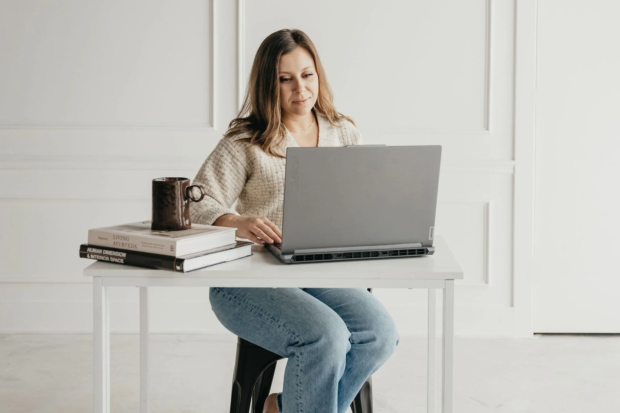 Woman sitting at a white desk working on a laptop with a stack of books and a cup nearby, in a bright room with white walls.