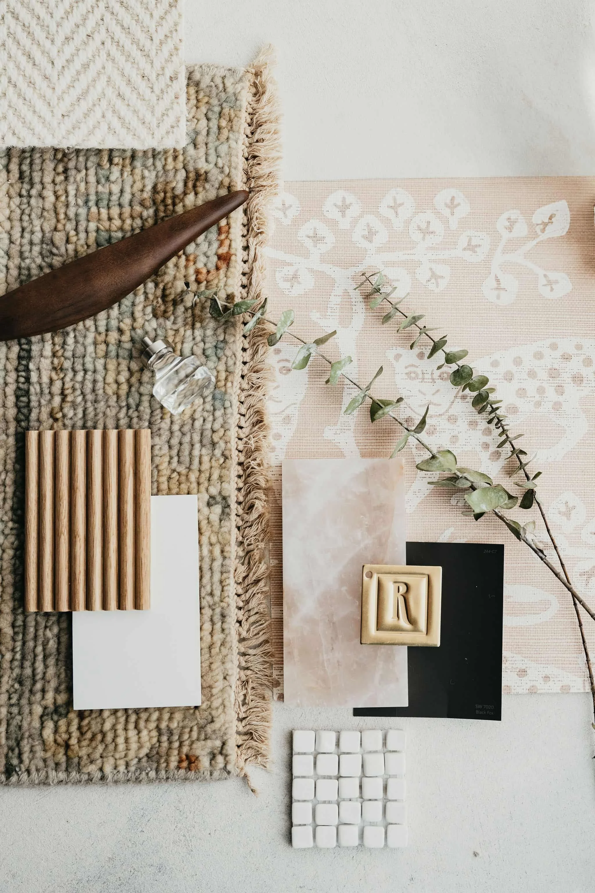 Flat lay of interior design and decor items on a neutral surface, including a textured rug, a wooden sculpture, a glass bottle, a leafy branch, a pink and cream patterned fabric, a marble tile, a cream decorative block with the letter R, a black sheet, a white grid, and small white tiles.