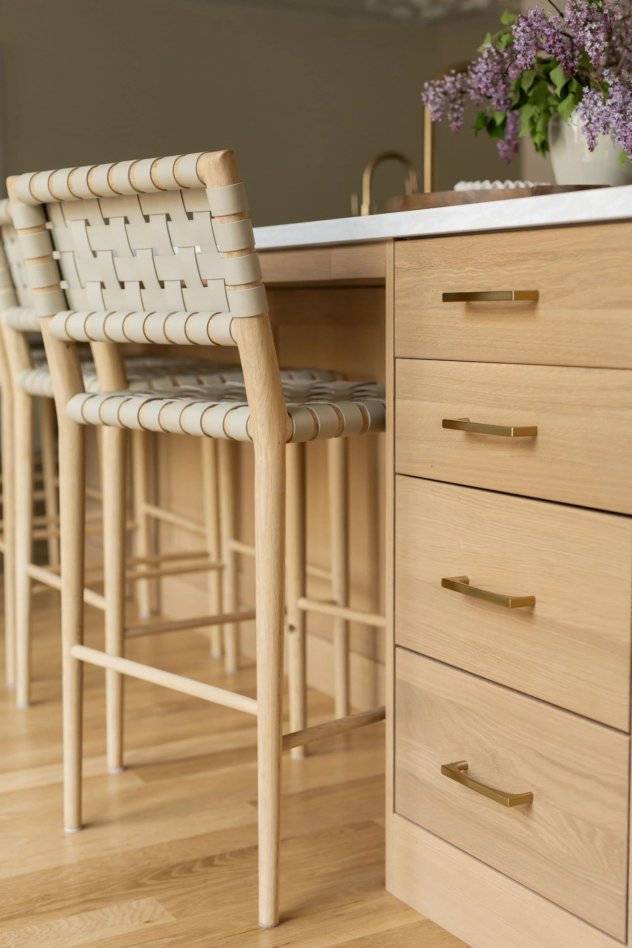 Close-up of a kitchen counter with a large vase of purple flowers, light wood cabinets with gold handles, and a bar stool with a woven seat.